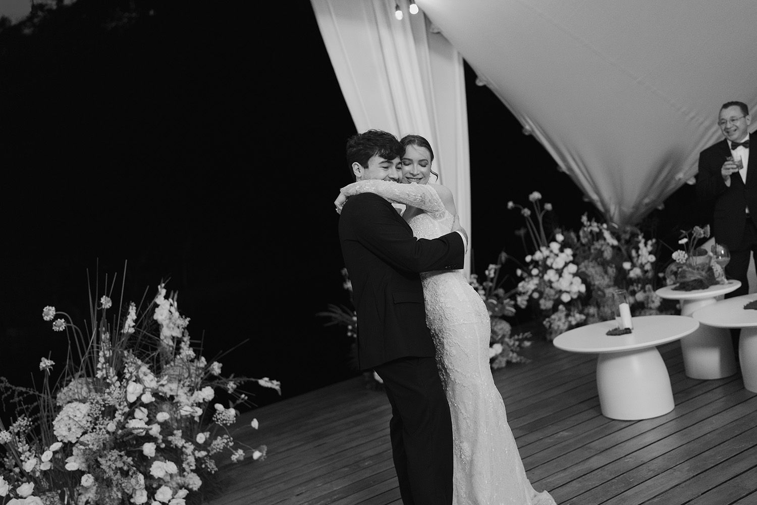 Black-and-white photograph of a bride and groom embracing as they dance beneath white drapery at their wedding reception.