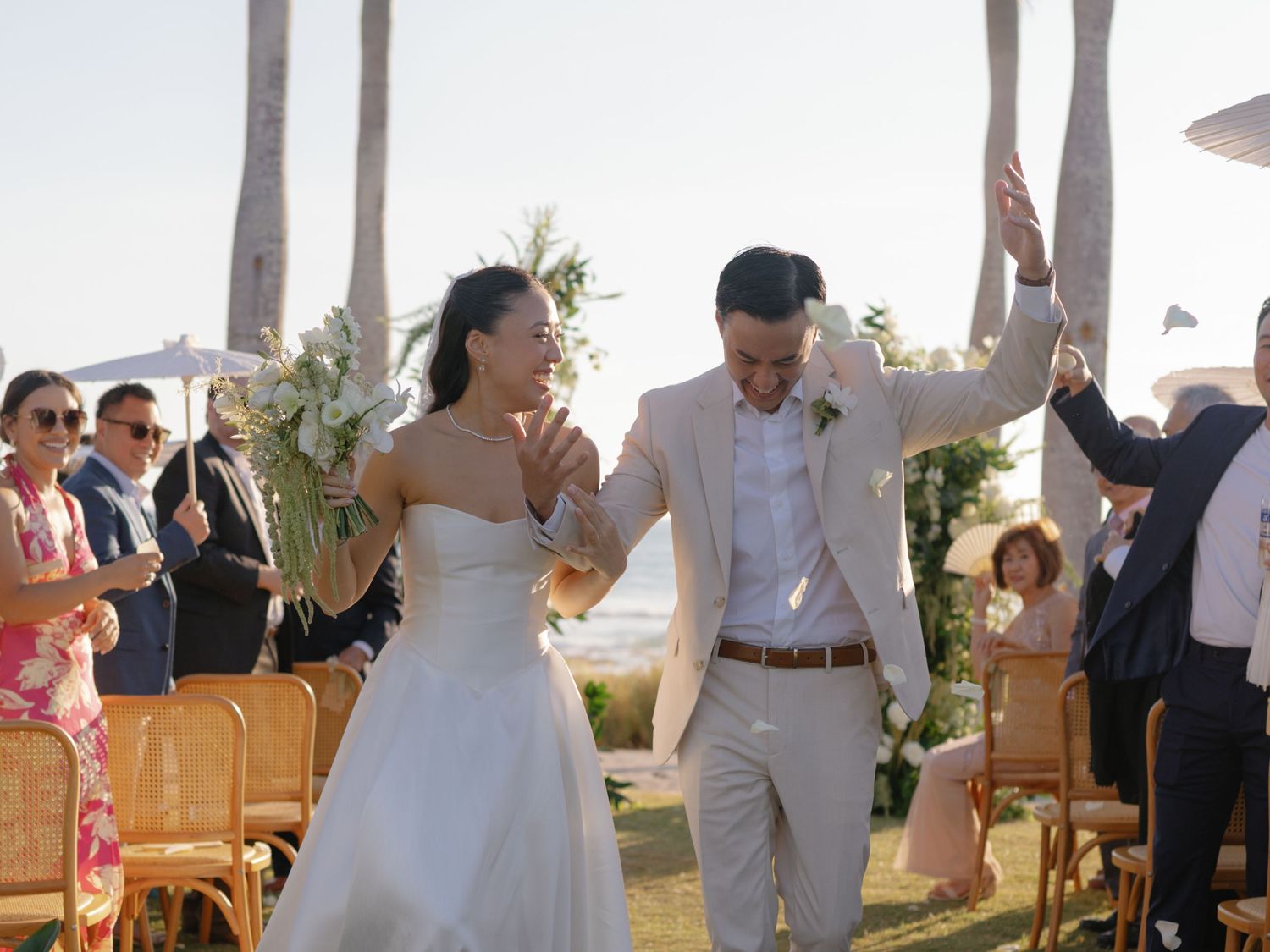 Joyful newlyweds walk down the aisle at an outdoor beach wedding ceremony, surrounded by guests and palm trees.