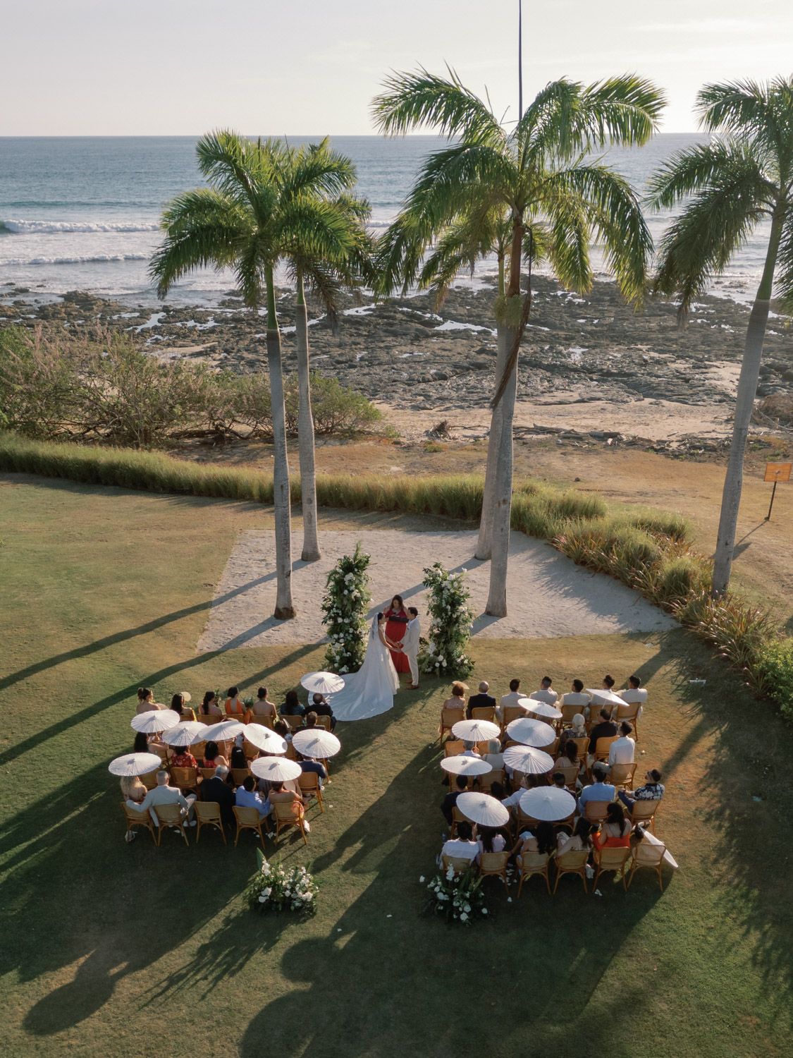 Oceanfront wedding ceremony in Costa Rica, with guests seated on a lawn beneath palm trees.