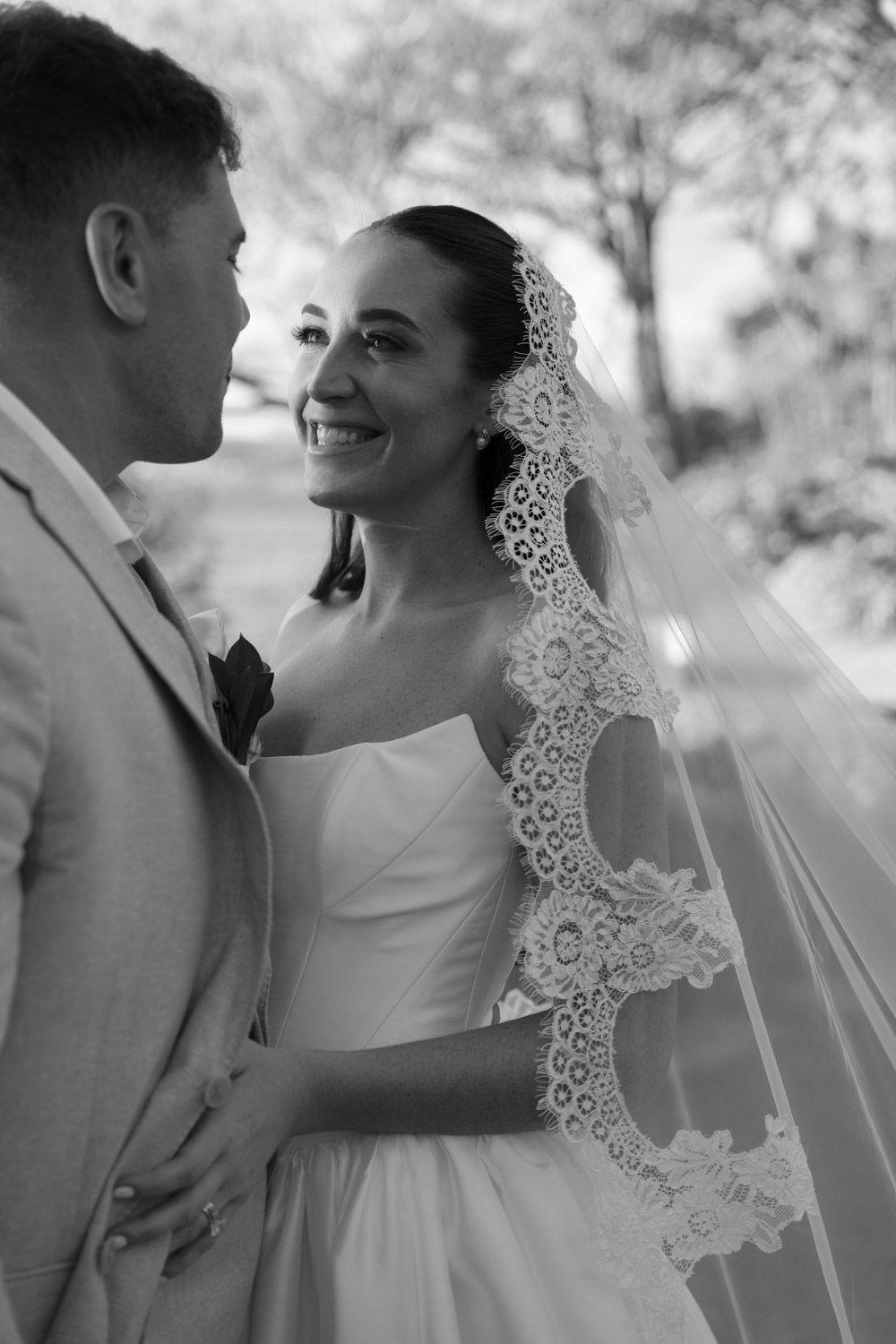 Black and white wedding photo of smiling bride in lace veil gazing at groom in light suit outdoors in costa rica