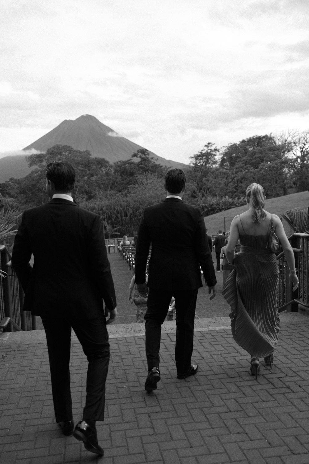Black and white photo of two men in suits and a woman in a gown walking toward a arenal wedding ceremony