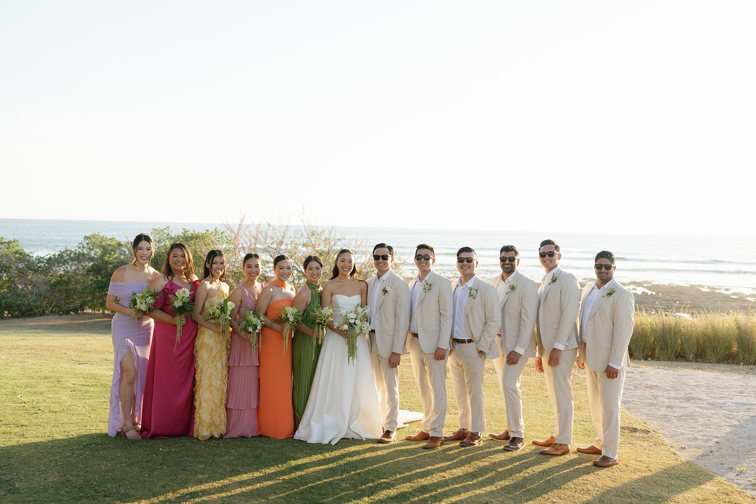 Bride and groom with bridesmaids in colorful dresses and groomsmen in beige suits, posing on an oceanfront lawn in Costa Rica.