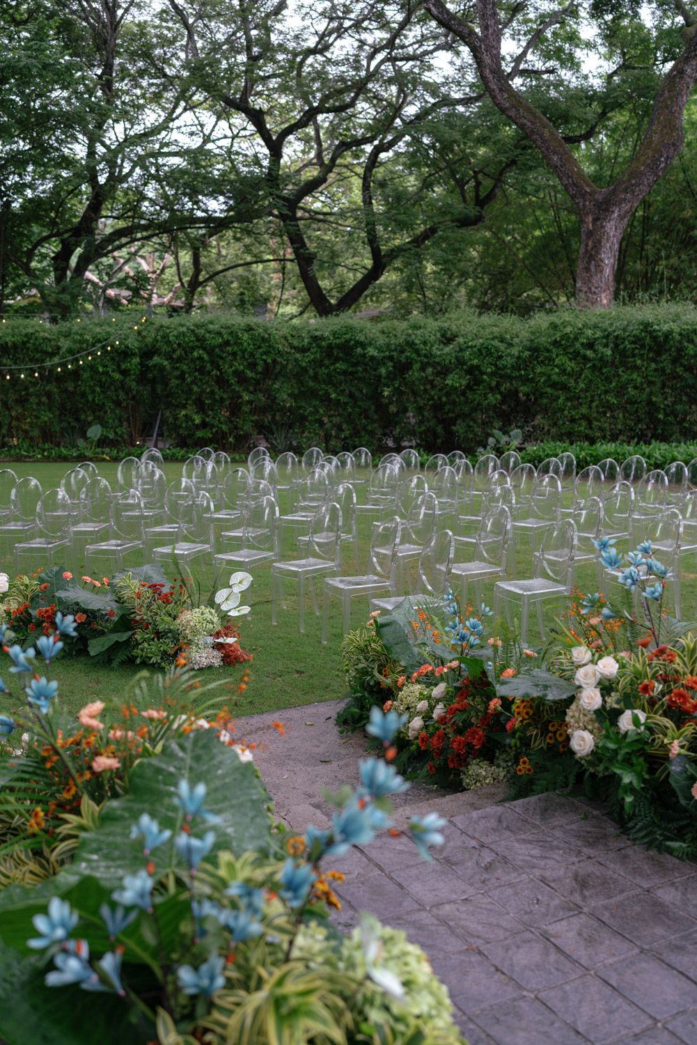 Outdoor wedding ceremony setup with clear acrylic chairs on green lawn, colorful floral arrangements in El Mangroove