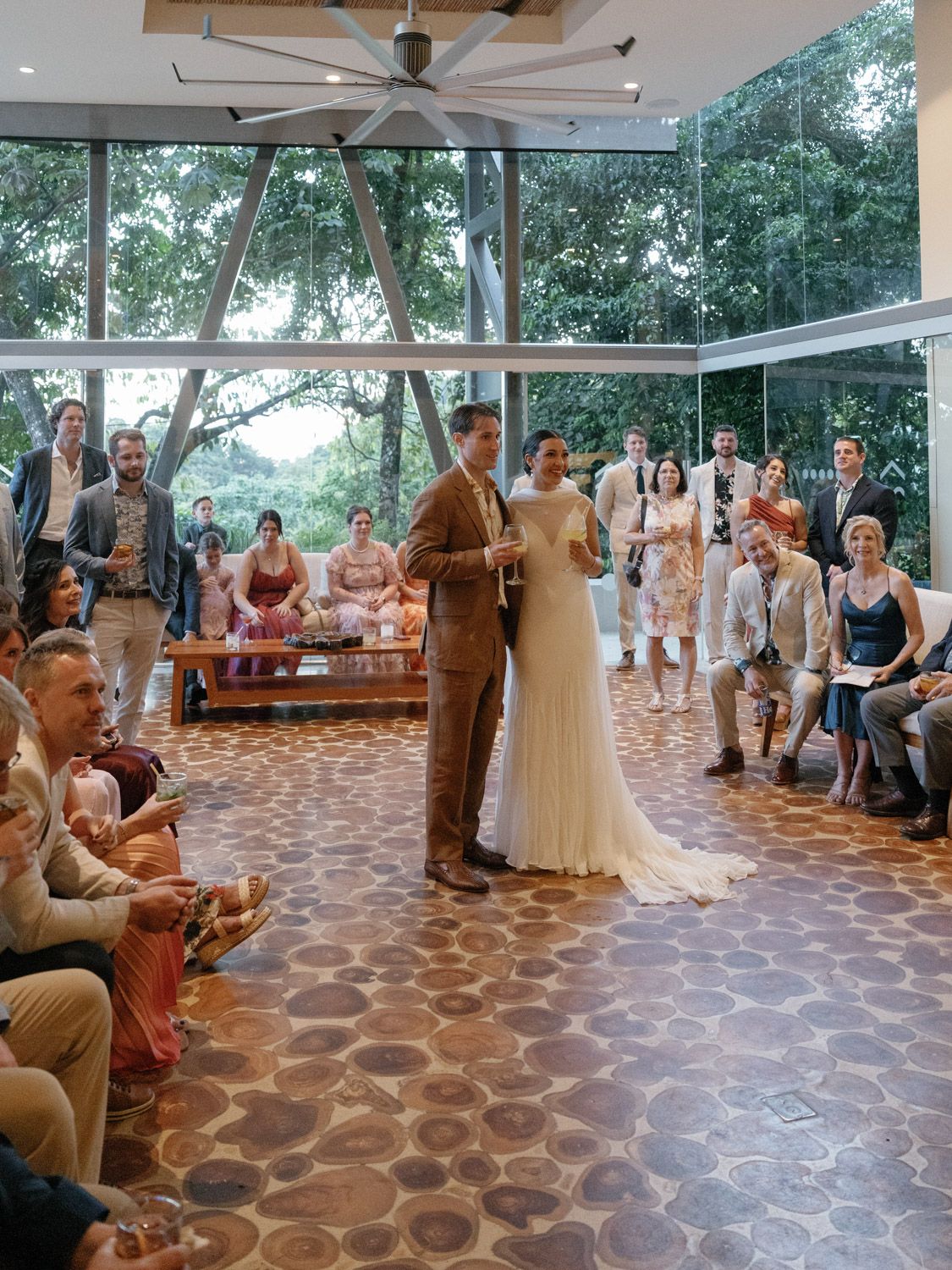 Couple exchanges vows during elegant indoor wedding ceremony with guests standing in glass-walled venue surrounded by lush greenery.