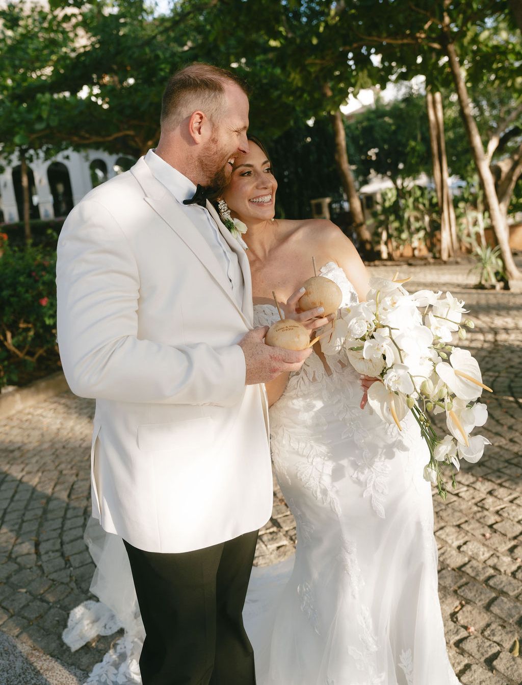 Groom in a white dinner jacket kisses smiling bride in a lace gown holding white flowers and coconuts in costa rica
