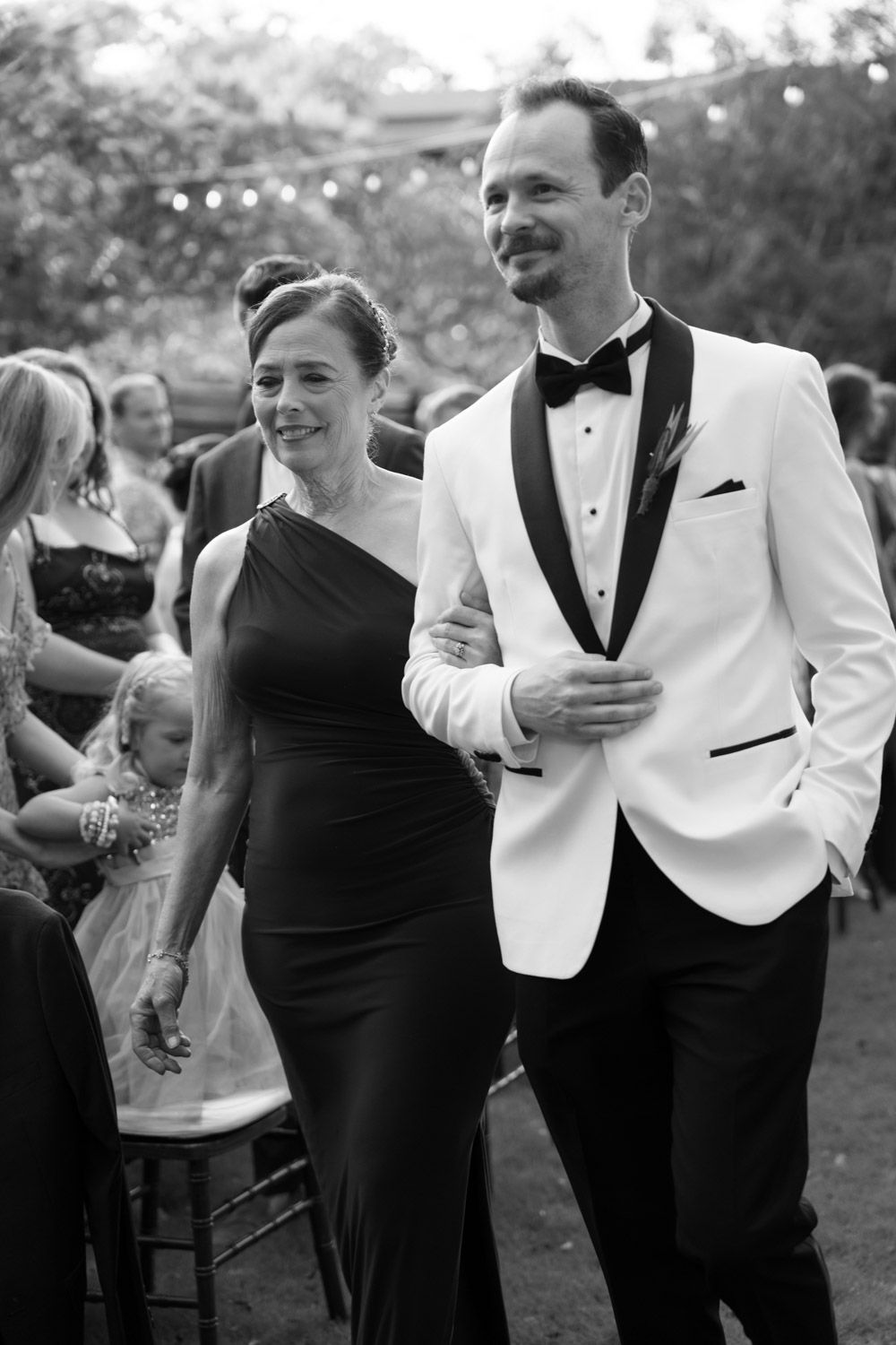 Black and white photo of a couple in formal attire walking at an outdoor wedding reception.