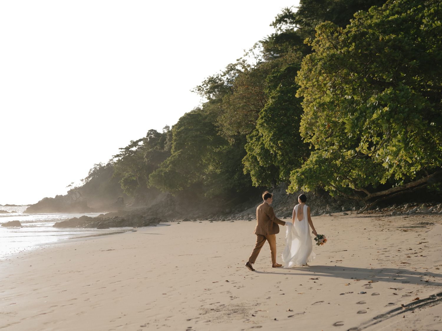 Wide view of Sadie and Ryan walking along a quiet beach backed by lush tropical trees.