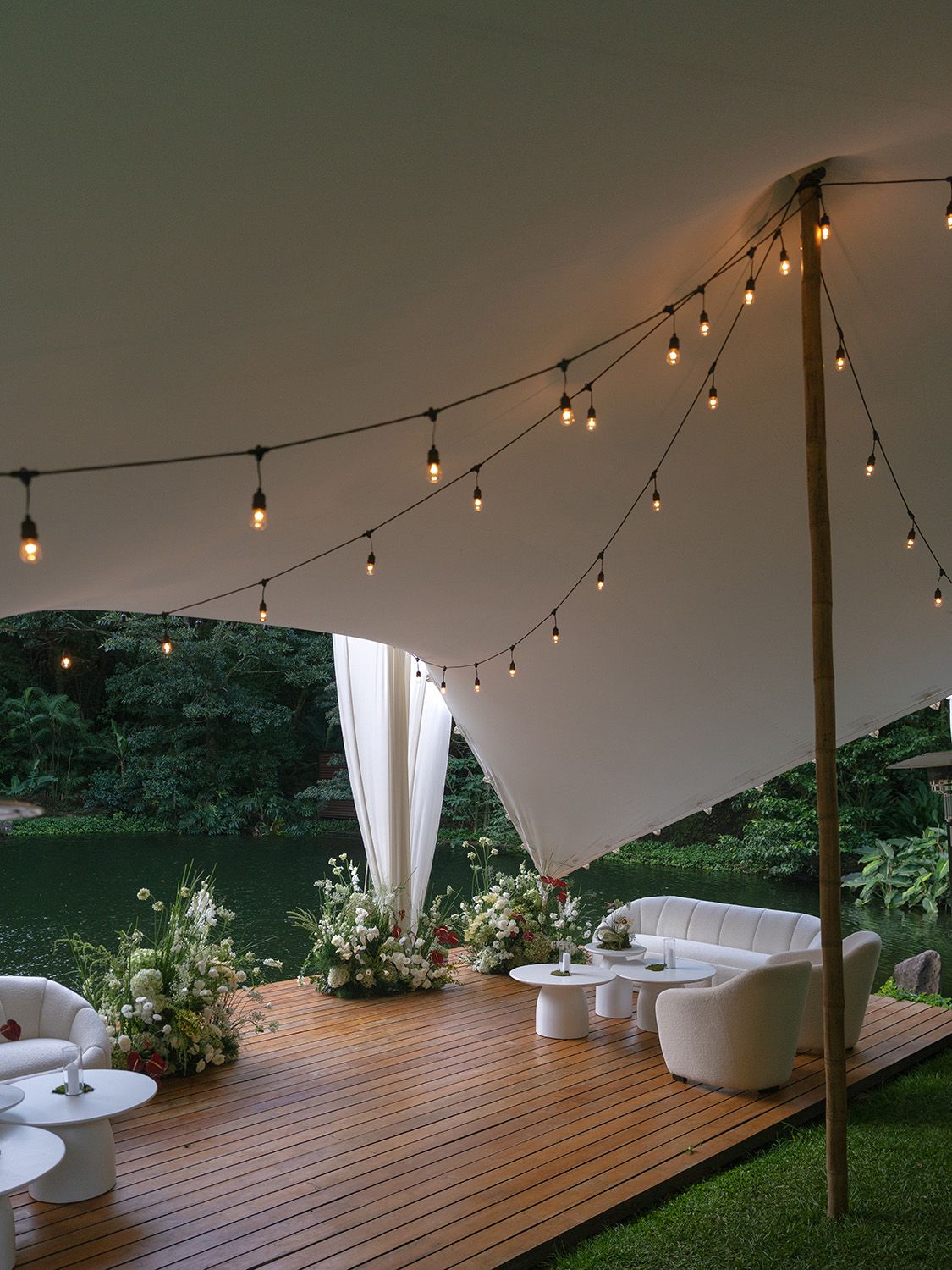 Wedding reception lounge in Costa Rica with white seating, floral arrangements, and string lights beside a pond.