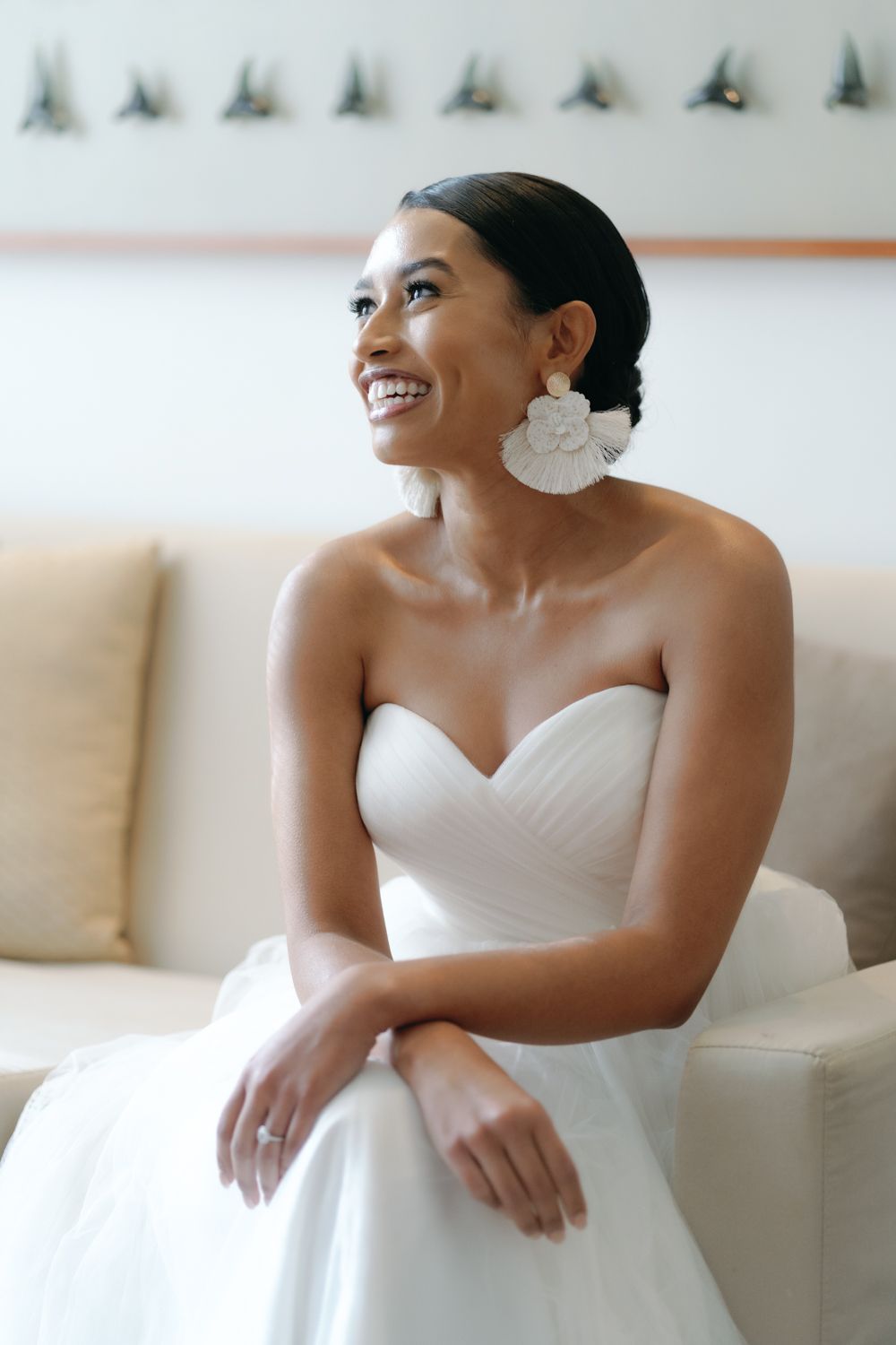 Smiling bride in a white strapless sweetheart wedding gown and large floral earrings sits on a cream sofa, gazing upward joyfully.