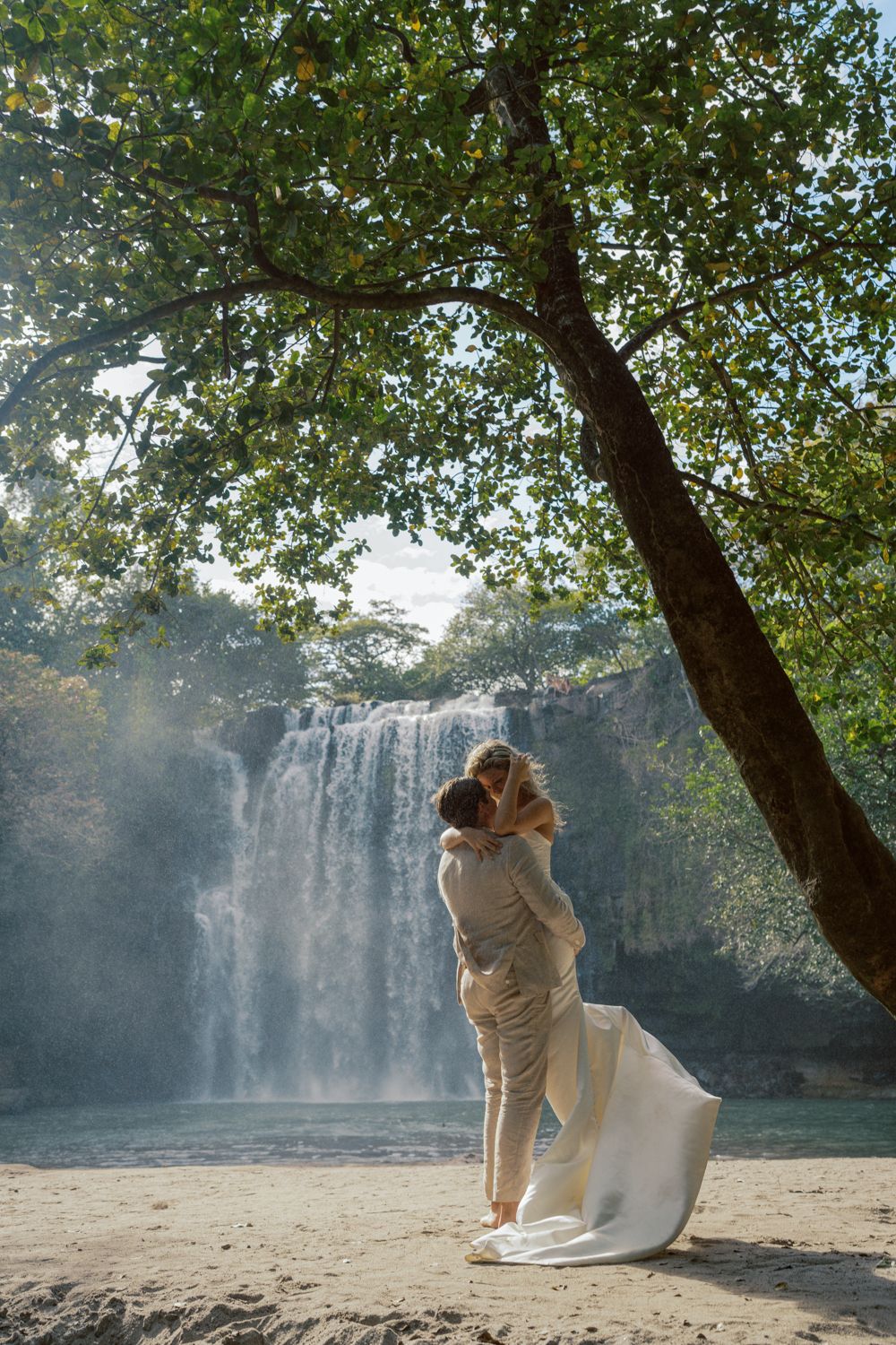 Groom lifting his bride beneath a leafy tree in front of a waterfall in Costa Rica.