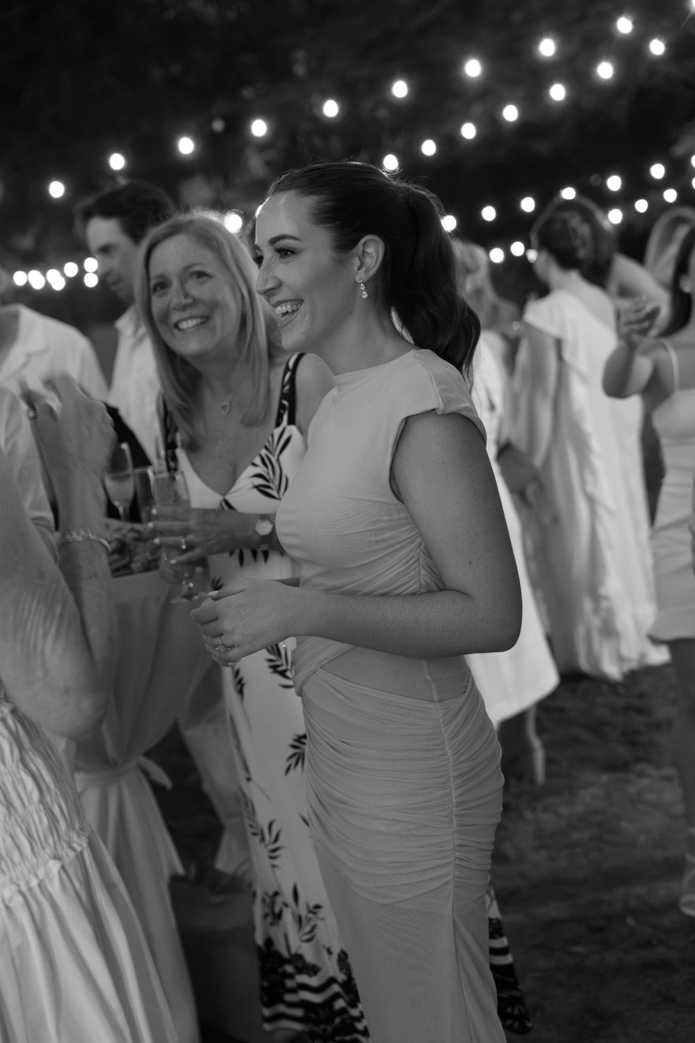 Black-and-white photo of guests laughing beneath string lights at a wedding welcome party in Costa Rica.
