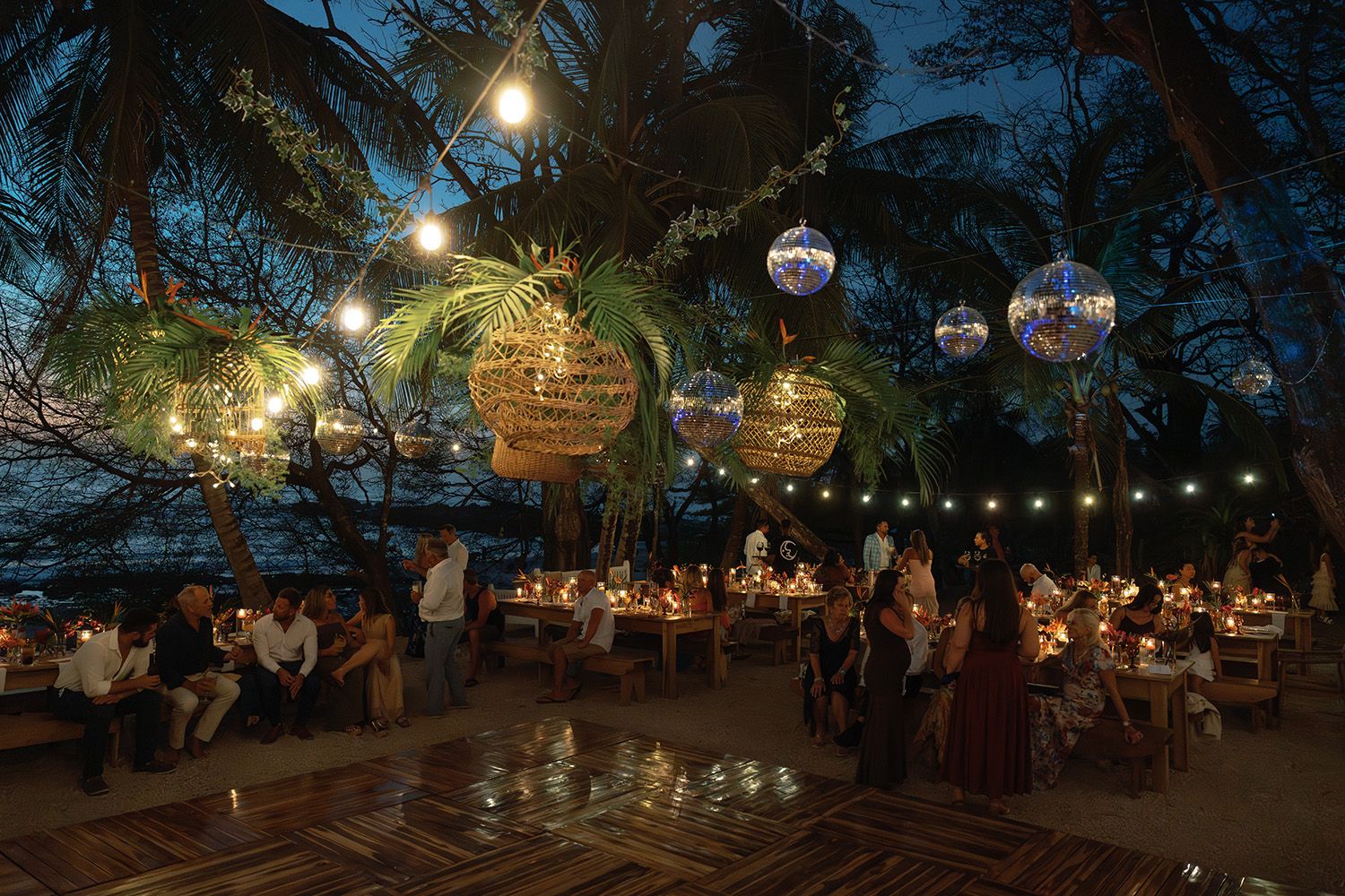 Beachside wedding reception at dusk with guests, string lights, woven lanterns, and disco balls beneath palm trees.