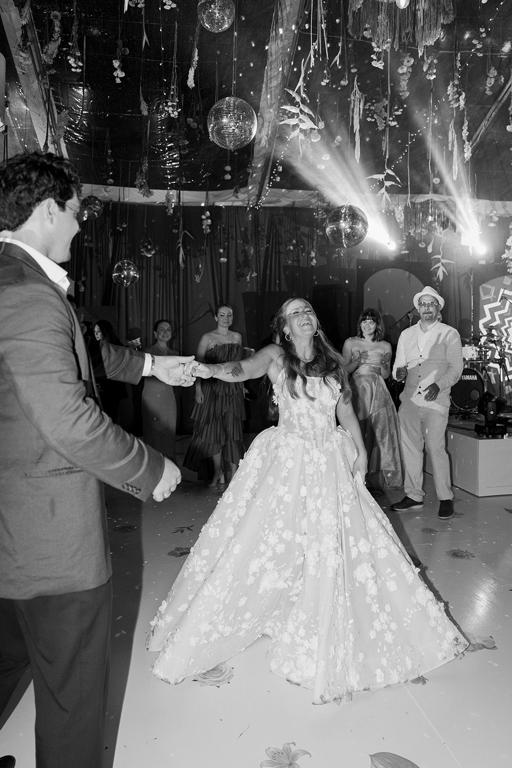 Black-and-white photograph of a bride laughing while dancing with the groom, surrounded by wedding guests beneath disco balls in Costa Rica.