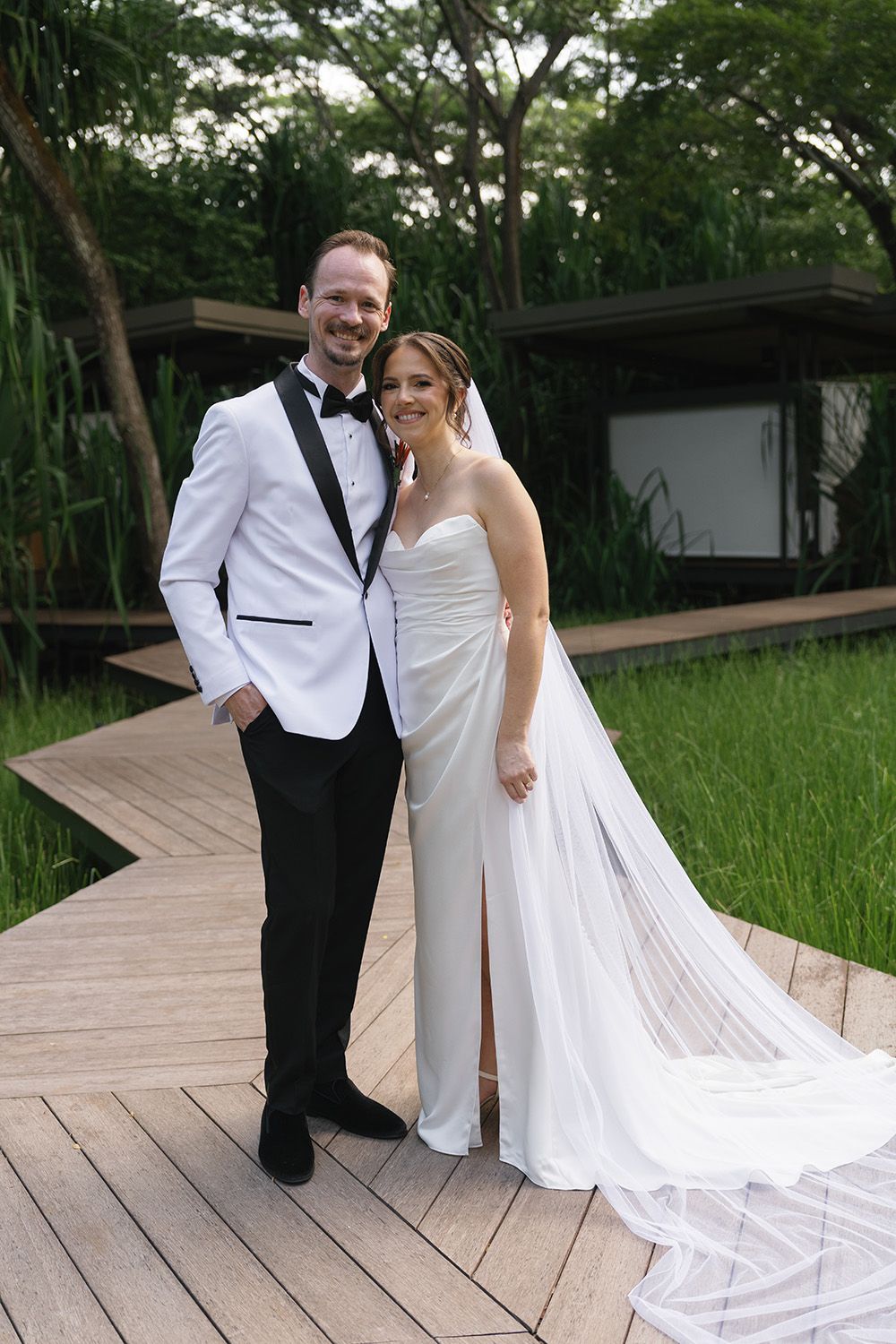 Bride and groom smiling together on a wooden walkway in a tropical garden in Costa Rica, with her long veil trailing behind.
