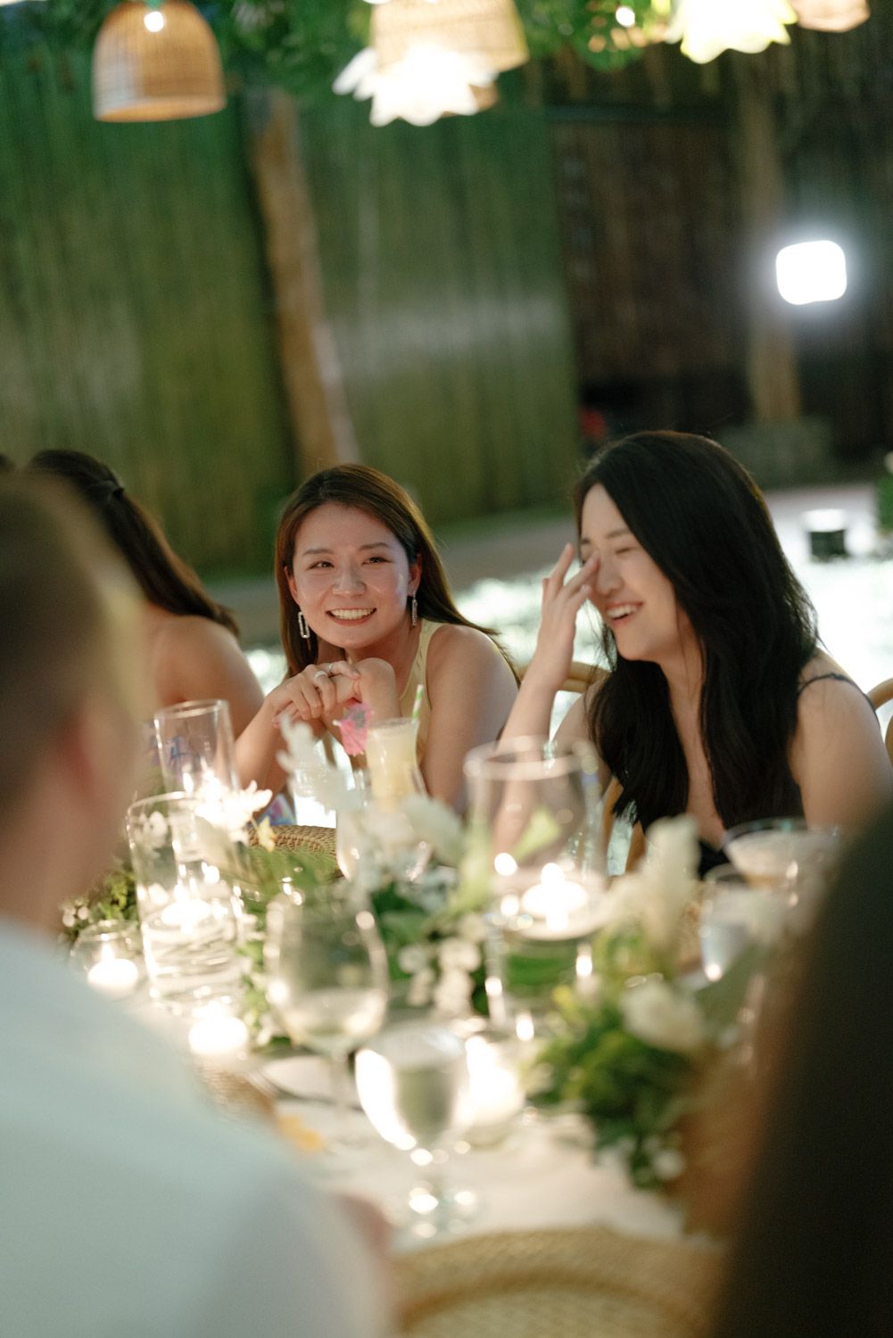 Two smiling guests laugh together at a candlelit outdoor dinner table with floral centerpieces, wine glasses, and warm string lights.