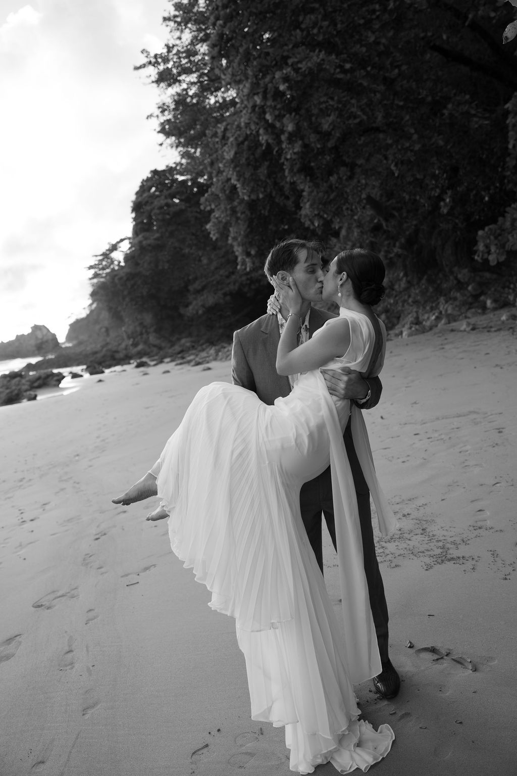 Black-and-white portrait of the groom holding and kissing the bride on the beach.