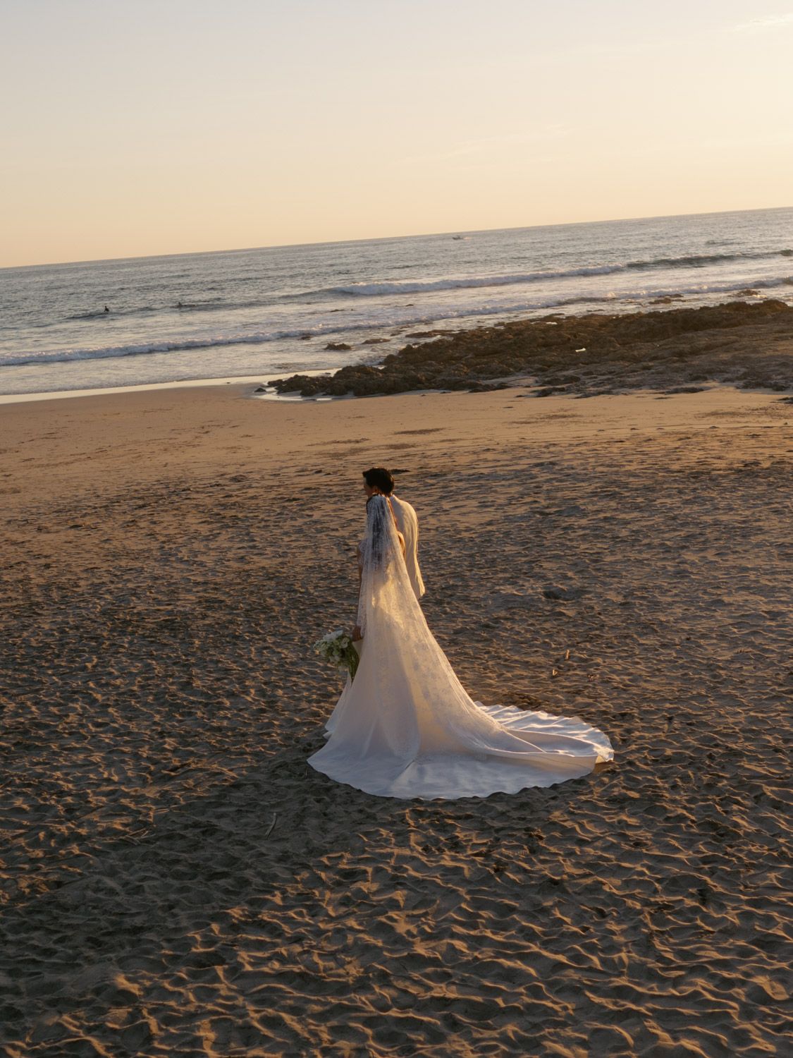 Bride in white wedding gown with long train walks on sandy beach at sunset with ocean waves in background in Costa Rica