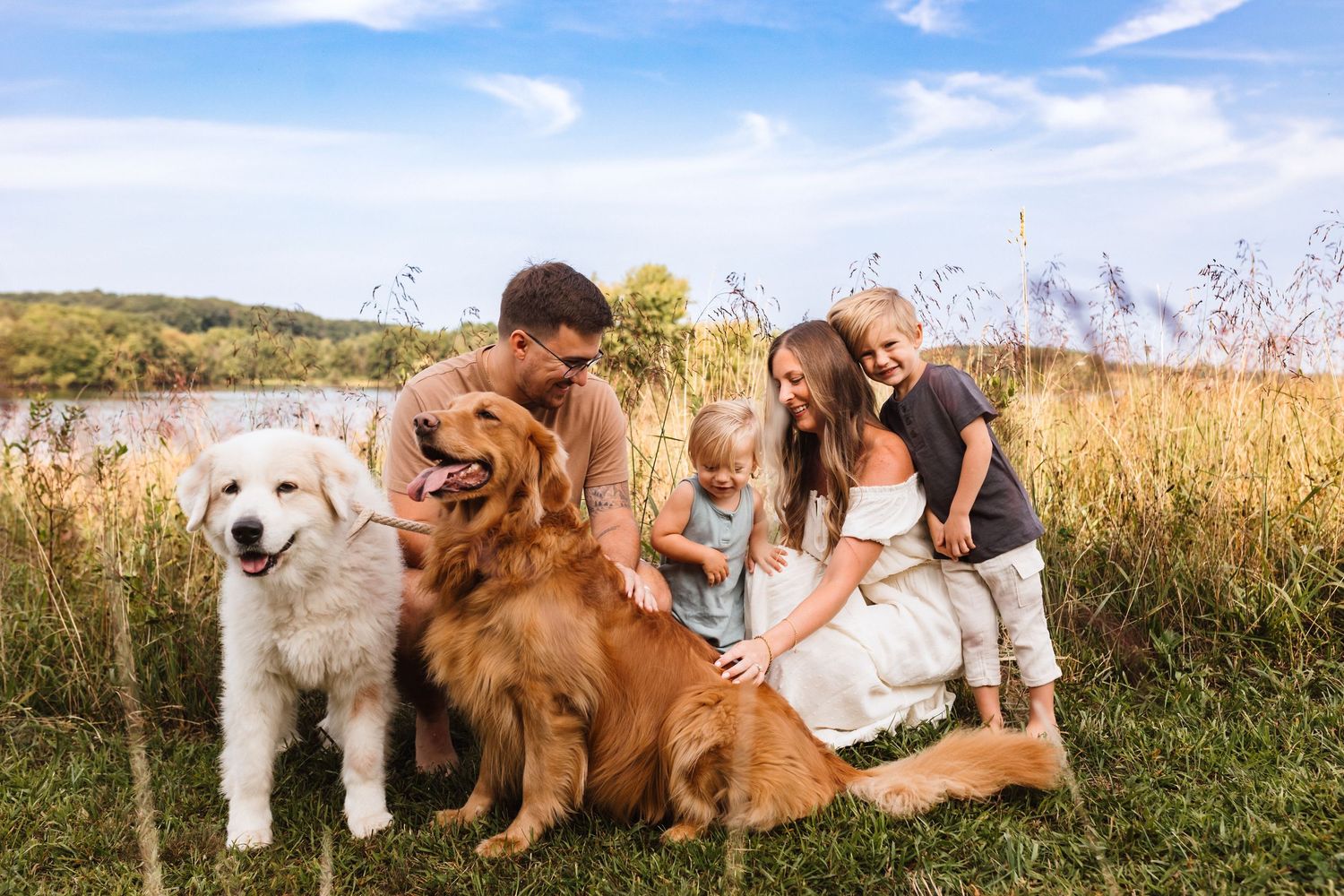 Happy family with two golden dogs posing near lake in tall grass on cloudy day.