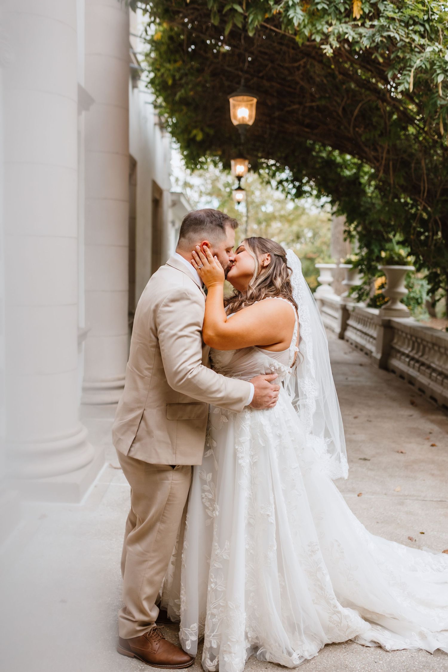 Bride and groom sharing a passionate kiss under a vine-covered archway with glowing lanterns at wedding.