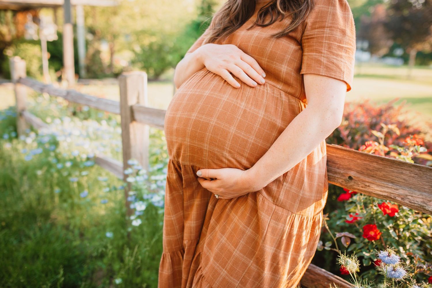 Pregnant woman in a rust plaid dress cradling her baby bump in a sunny garden setting.