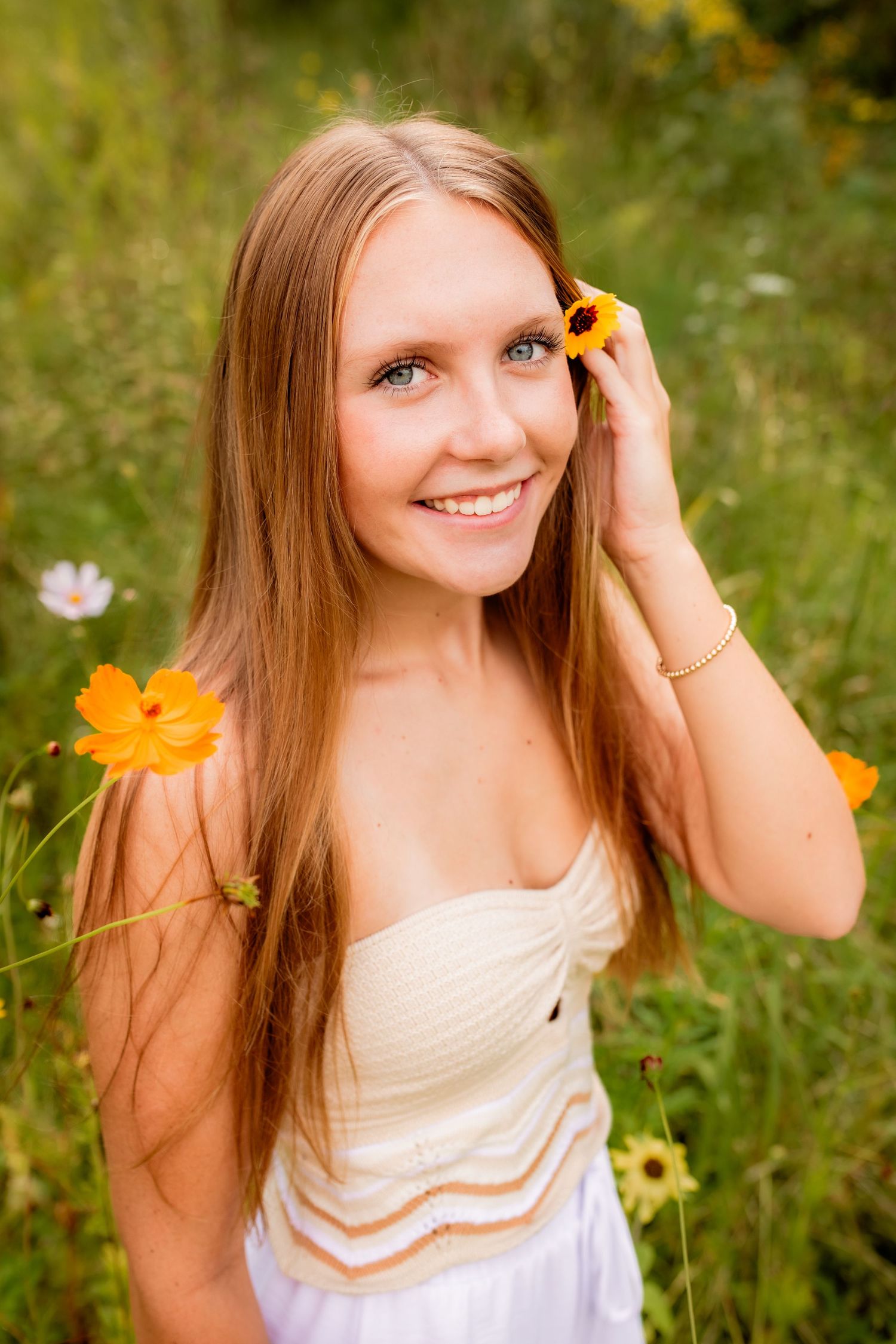 Smiling blue-eyed teen girl holds yellow cosmos flower near her ear in a vibrant wildflower field.