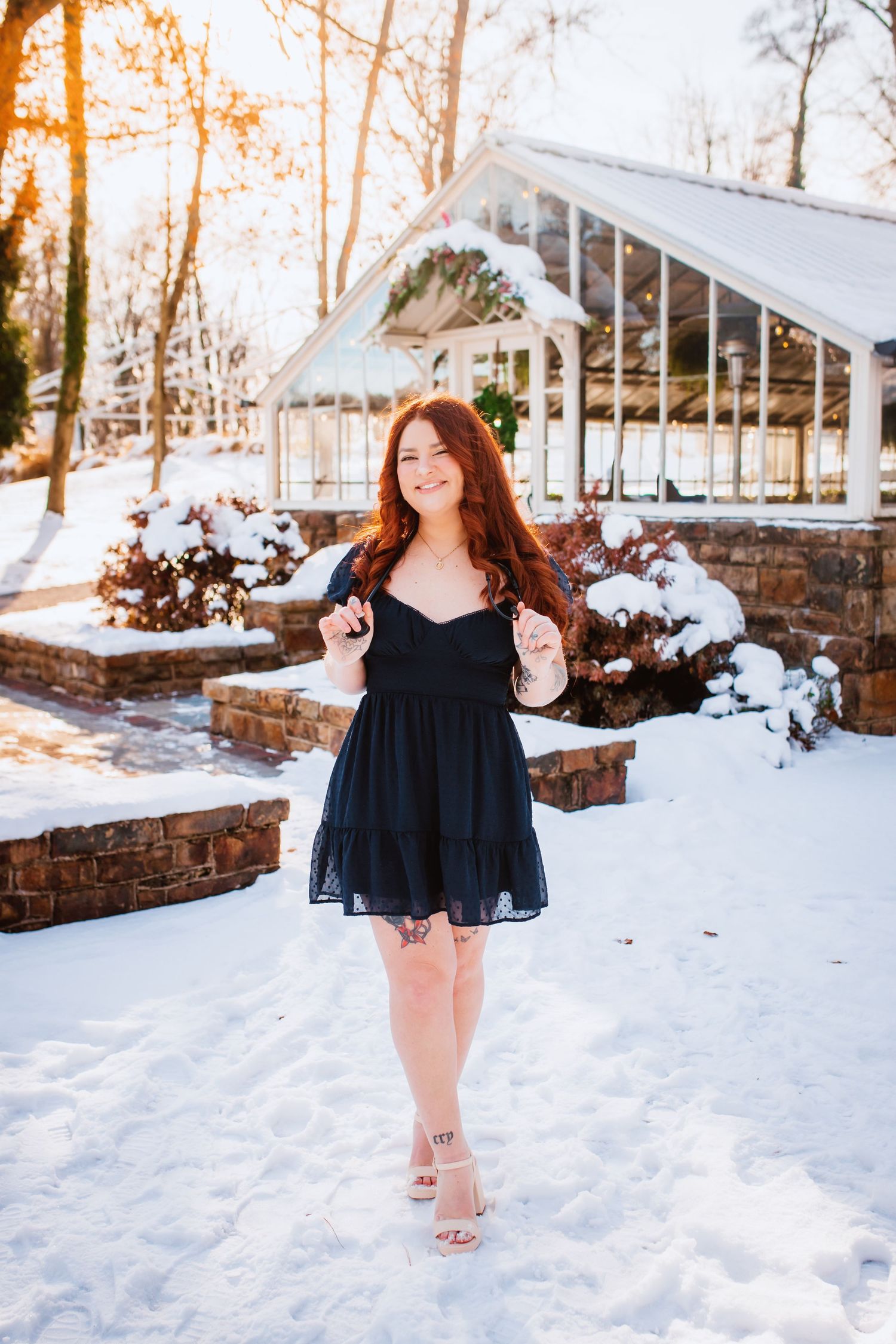 Redhead woman in black mini dress poses in snowy outdoor venue at sunset.
