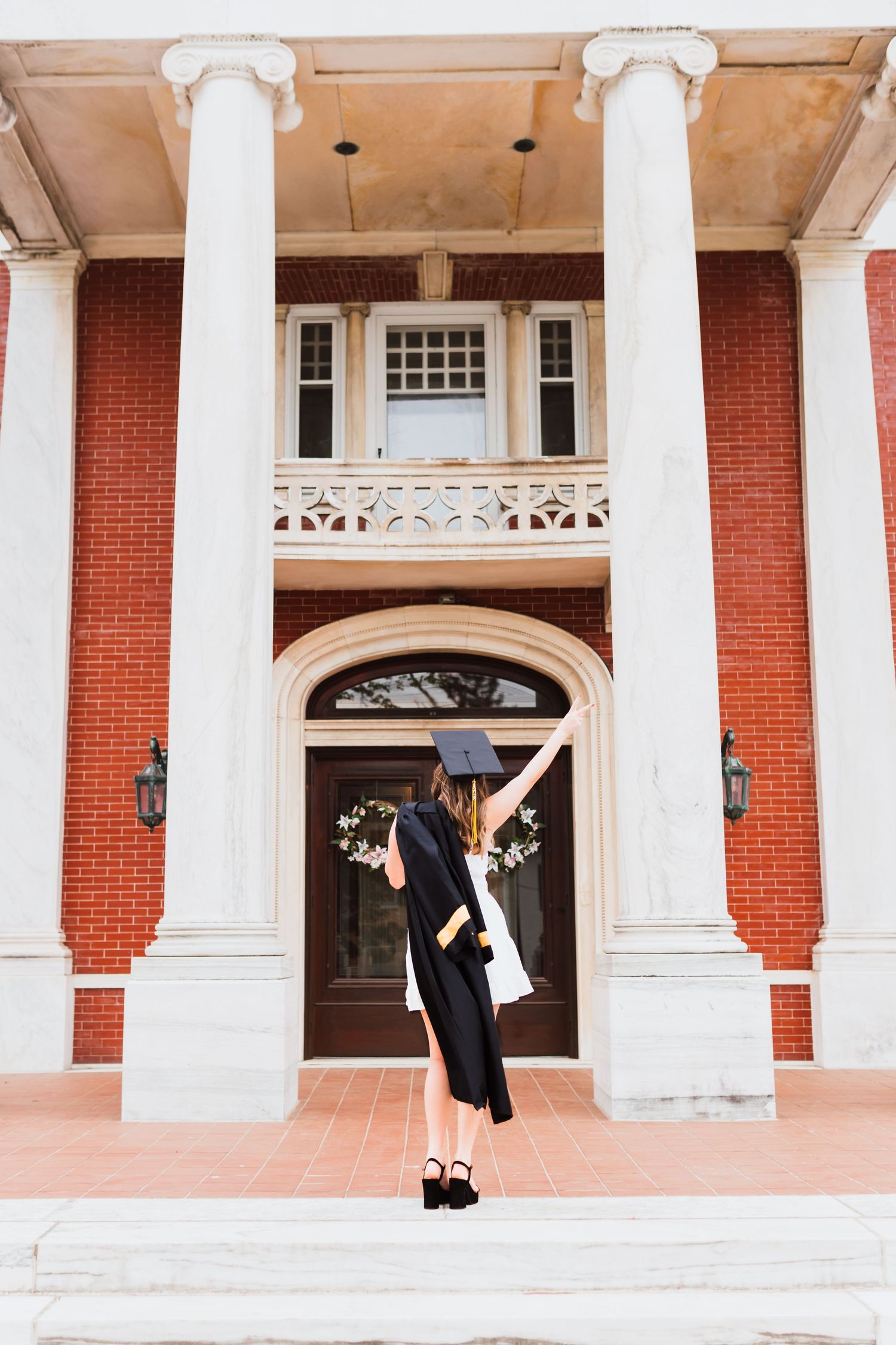 Graduate in black cap and gown raises arm triumphantly on steps of classic brick building with columns.