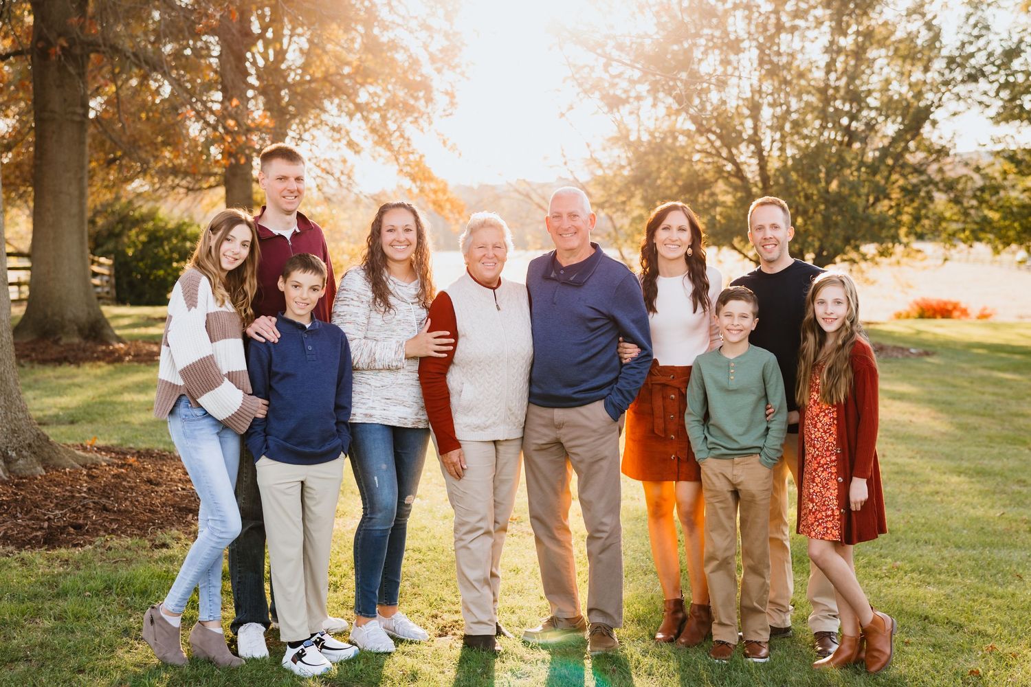 Extended family of nine posing together on green lawn with golden autumn trees glowing in background.