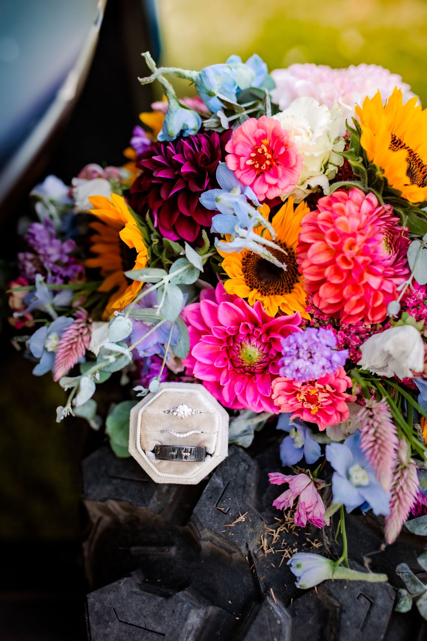 Vibrant wedding bouquet featuring sunflowers, dahlias, and mixed blooms with a ring box resting on rustic wood.