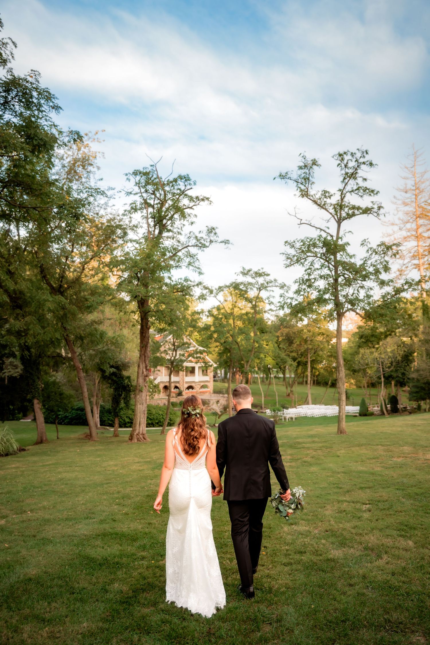 Bride and groom walk hand in hand across a lush green lawn toward a historic estate.