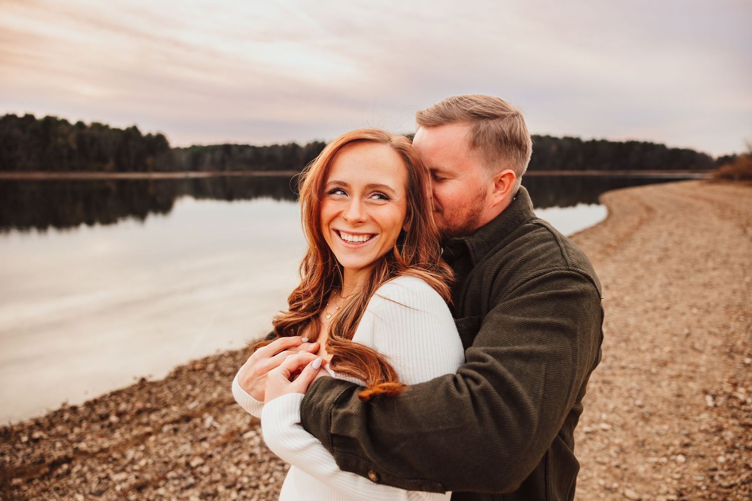 Happy couple sharing a romantic moment by a lake at sunset, woman smiling as man embraces her from behind.