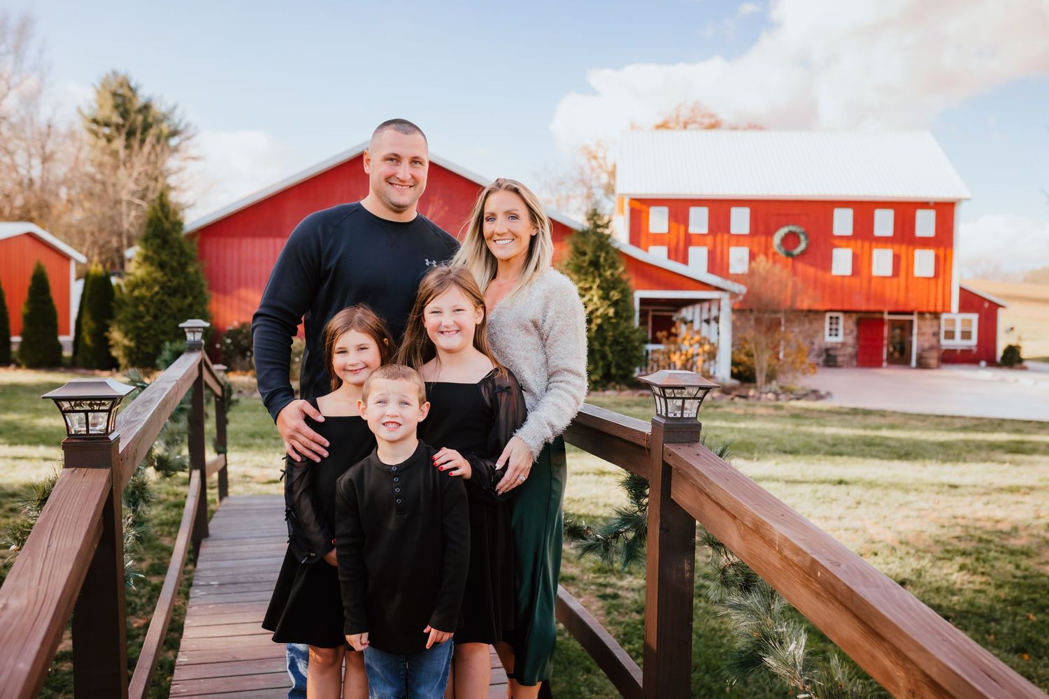 Family of five posing on wooden bridge with red barn backdrop during golden hour outdoor family portrait session.
