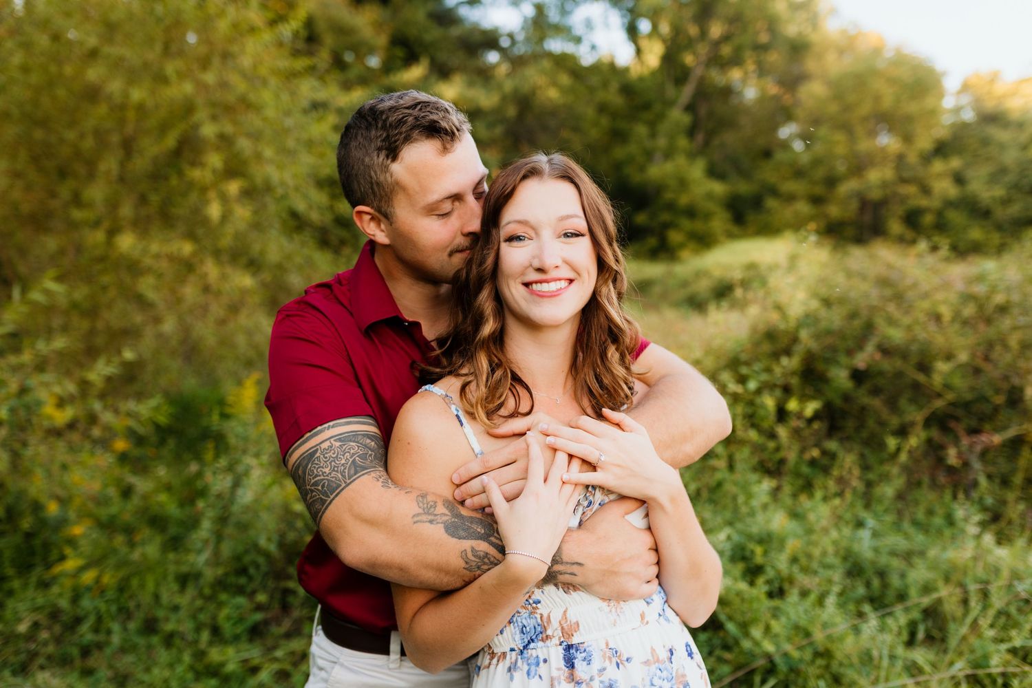 Tattooed man embracing smiling woman in floral dress in a green meadow.