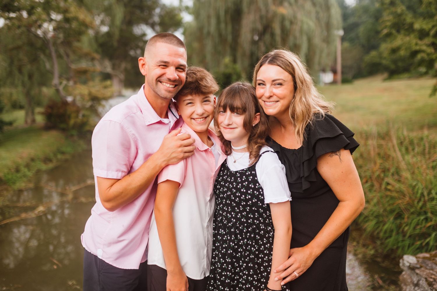 Smiling family of four poses together near a pond in an autumn park.