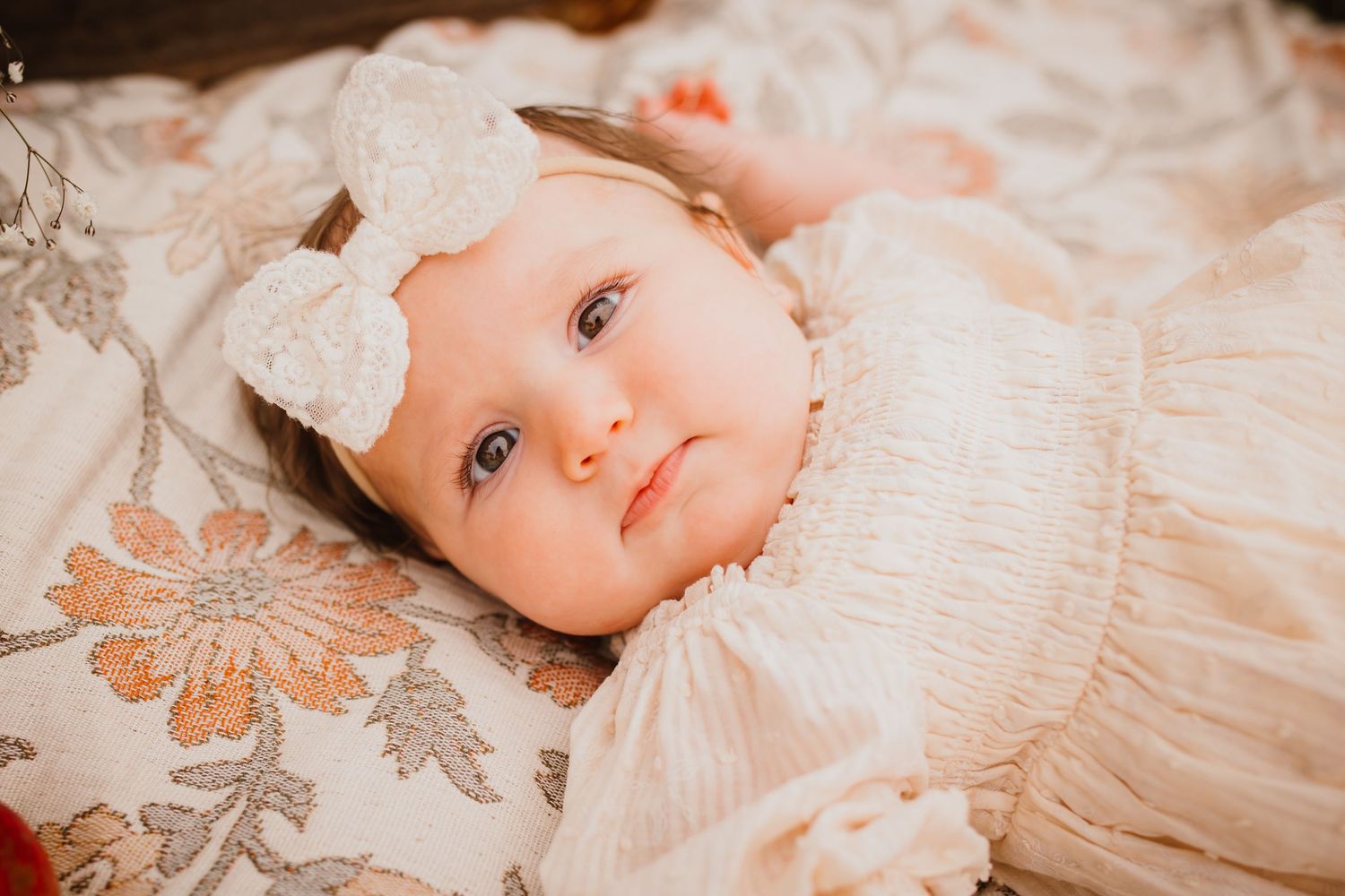 Adorable baby girl in a white lace dress and bow headband lying on a floral blanket.