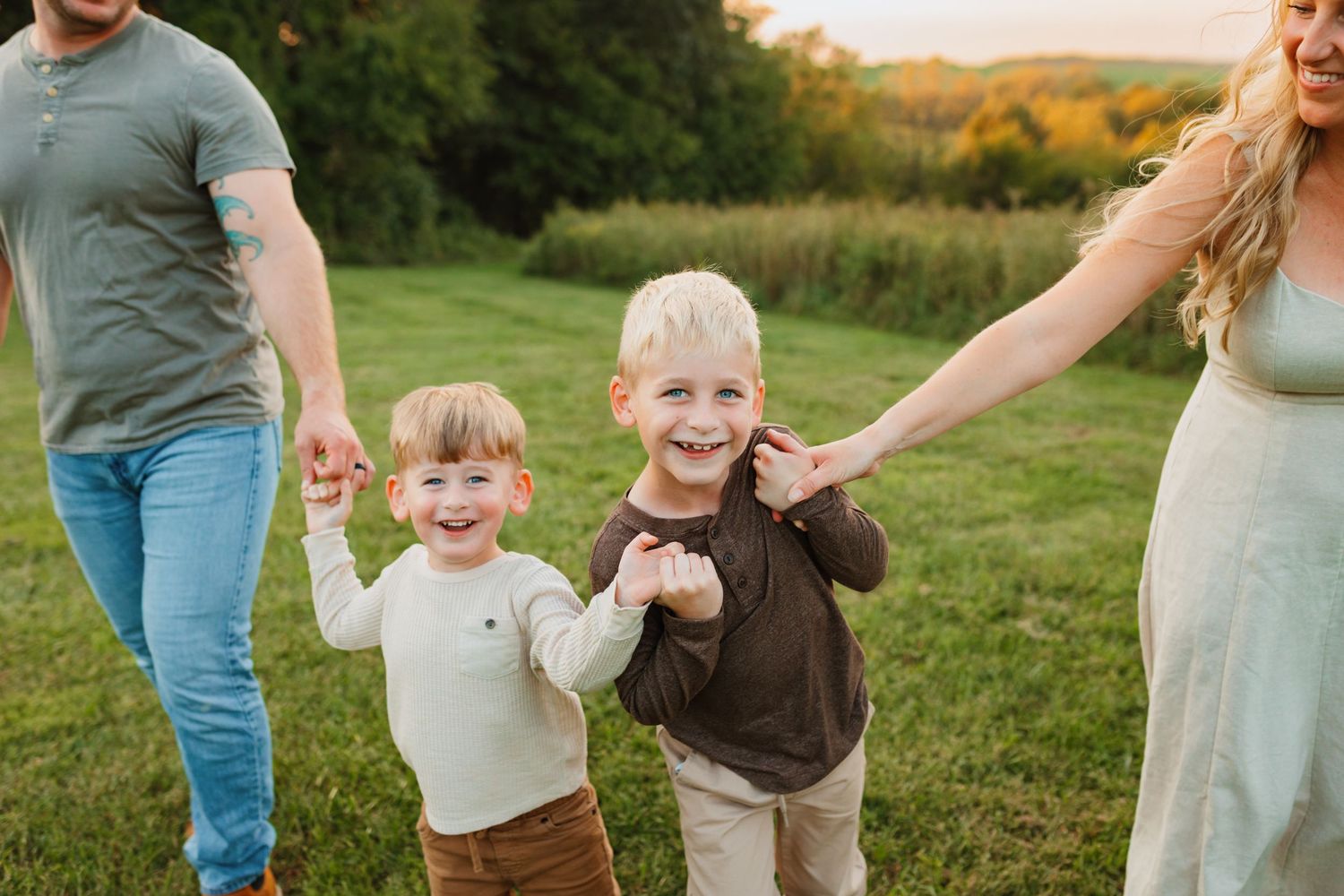 Two smiling young boys hold hands with their parents while walking across a sunny grassy field with trees in the background.