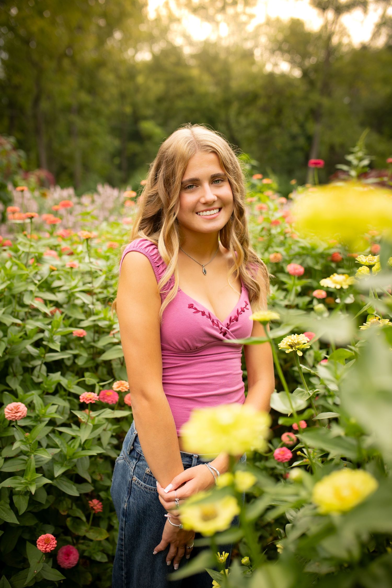 Smiling young woman in pink top standing among colorful zinnia flowers in a lush garden setting.