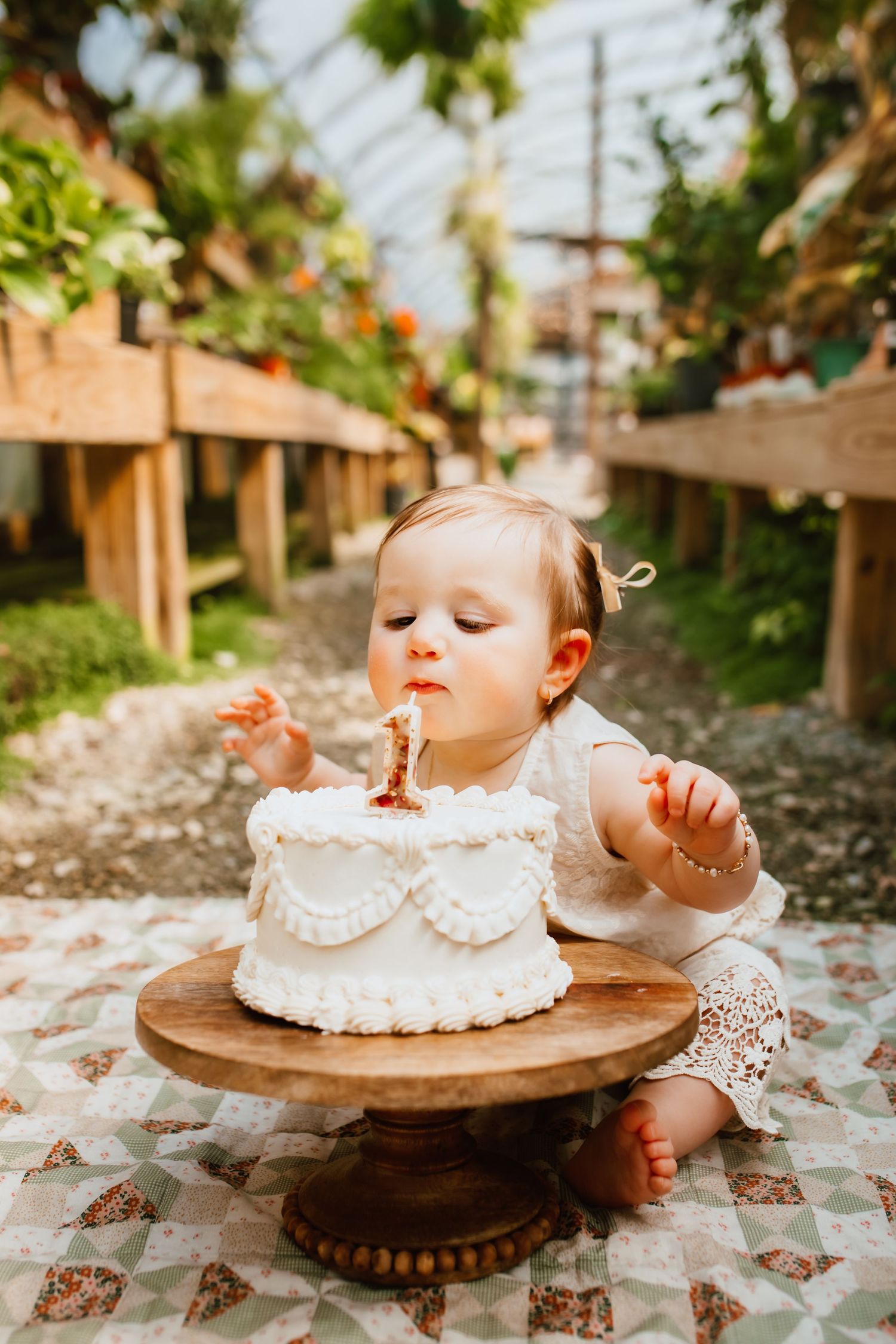 Baby girl in white lace dress blowing out candle on first birthday smash cake outdoors.