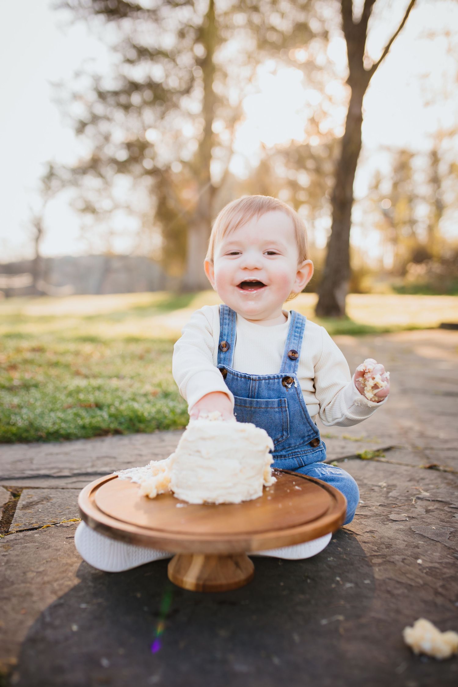 Joyful baby in denim overalls smashing a white cake on a wooden stand outdoors at golden hour.