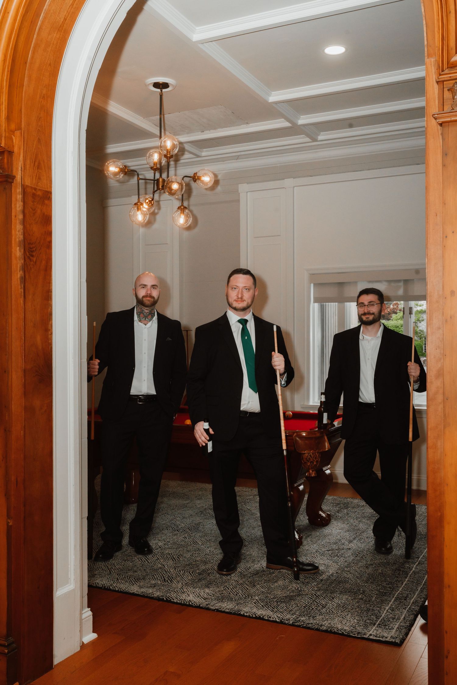 Three groomsmen in black suits standing under archway holding billiard cues in elegant home.