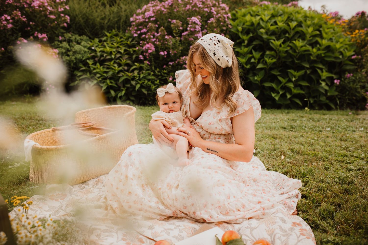 Mother in floral dress sitting on blanket in garden holding infant baby surrounded by pink blooming flowers.