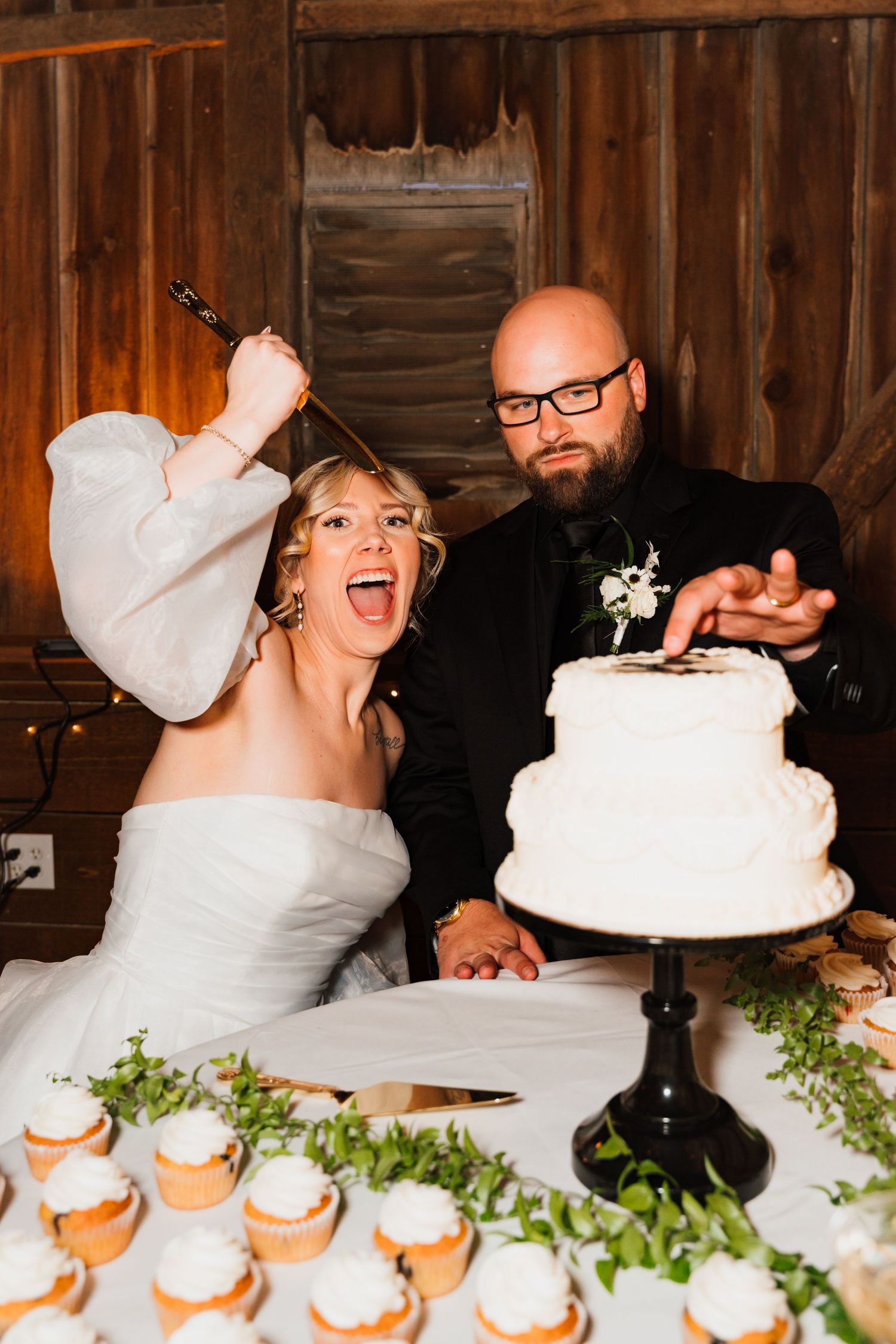 Excited bride and groom cut white two-tier wedding cake at rustic barn reception venue.