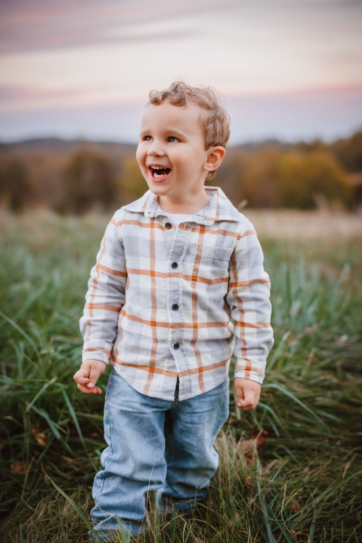 Happy toddler boy in plaid shirt and jeans laughs in an open grassy field at sunset.