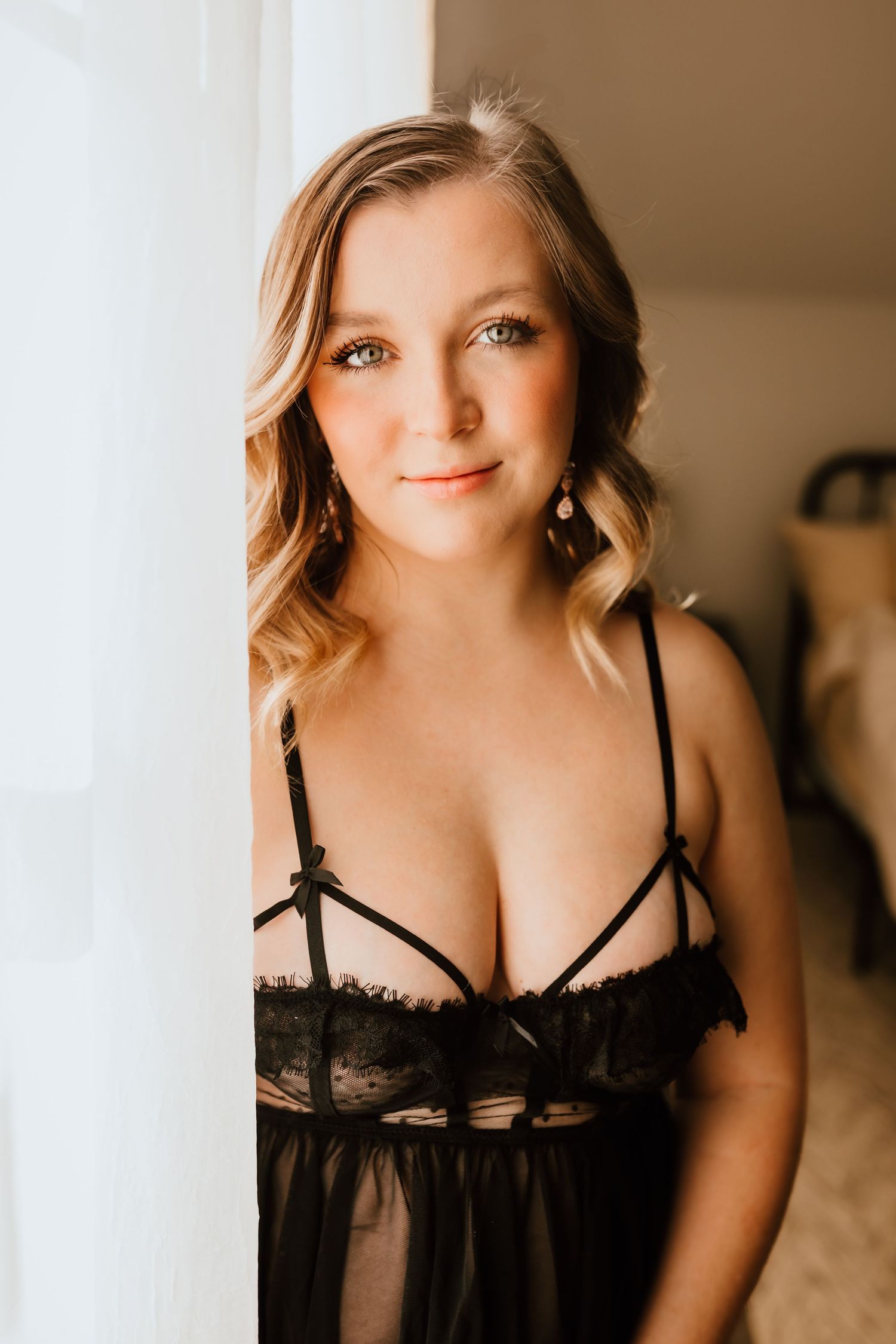 Blonde woman in black sheer lace lingerie stands by white curtain smiling.