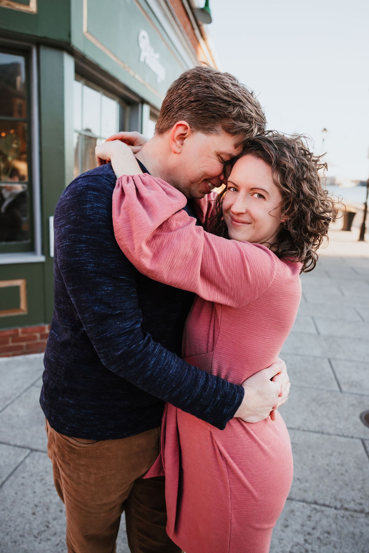 Couple sharing a warm embrace on a city sidewalk, woman in pink dress smiling while man holds her close.