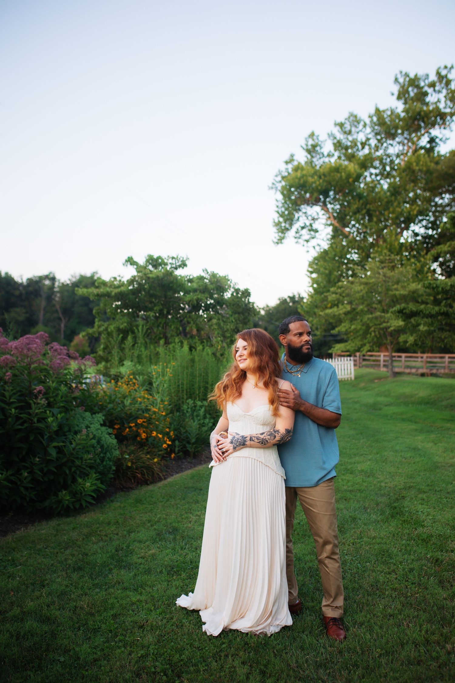 Tattooed interracial couple standing together in a lush garden during golden hour engagement shoot.