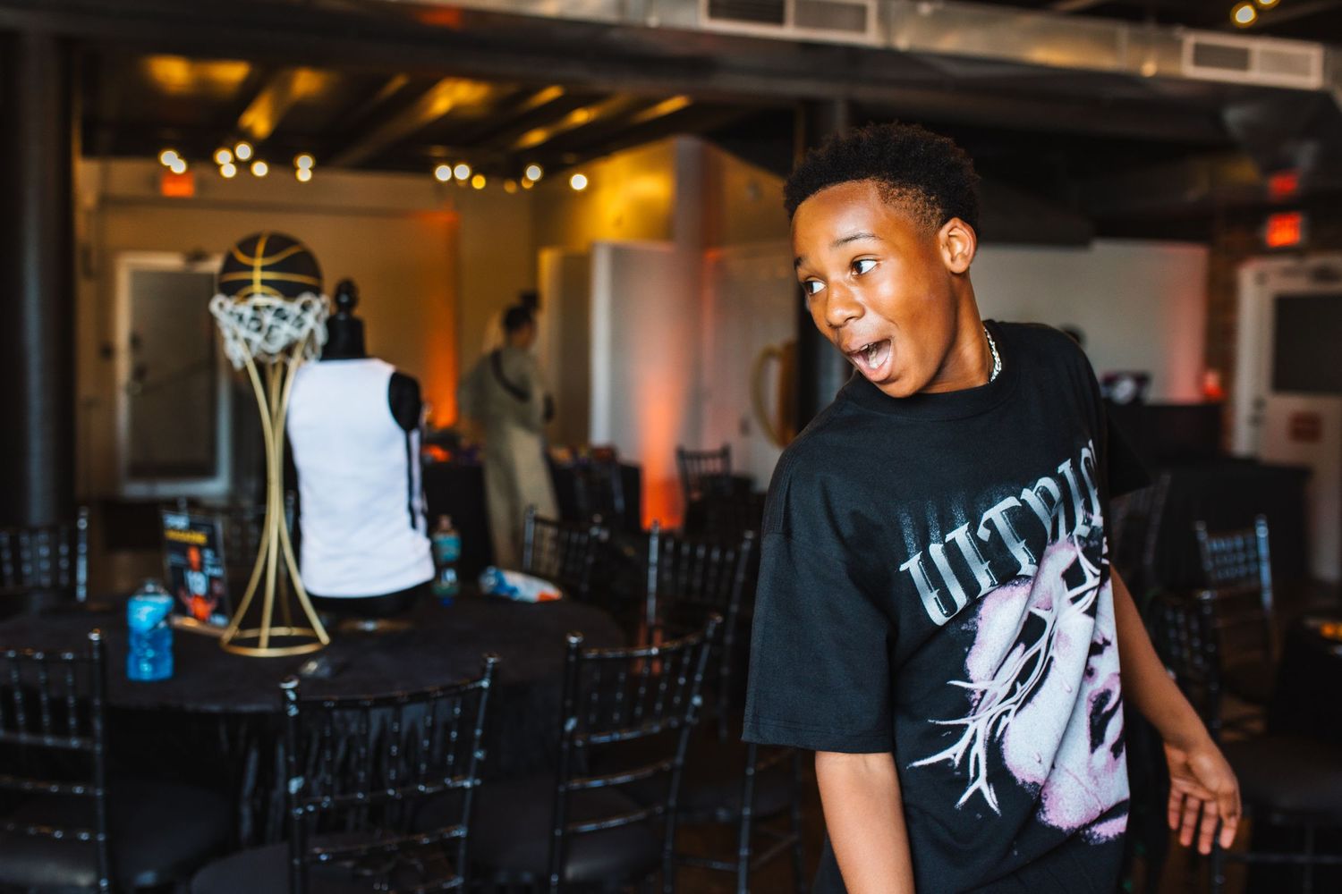Excited young Black boy in black graphic tee at basketball-themed birthday party celebration with trophy centerpiece.