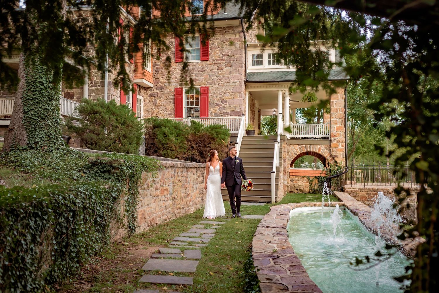 Bride and groom stroll along a stone path beside a fountain near an ivy-covered estate.