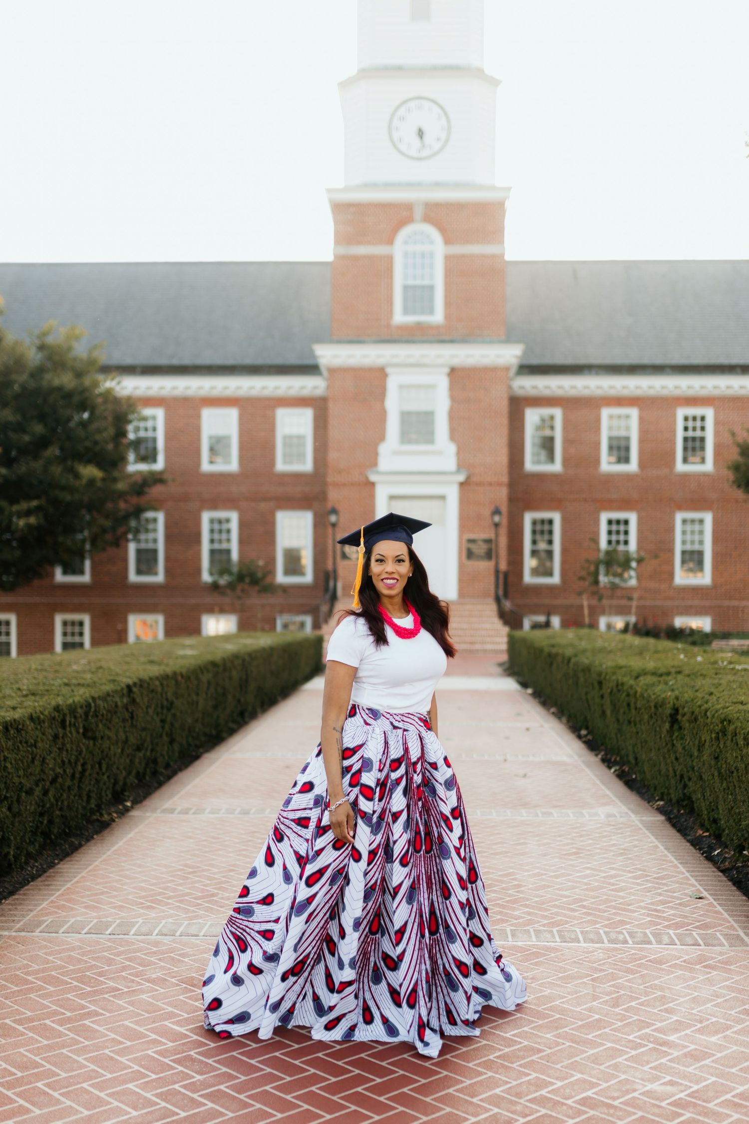 Graduate in black cap, white top, and colorful maxi skirt standing before brick building.