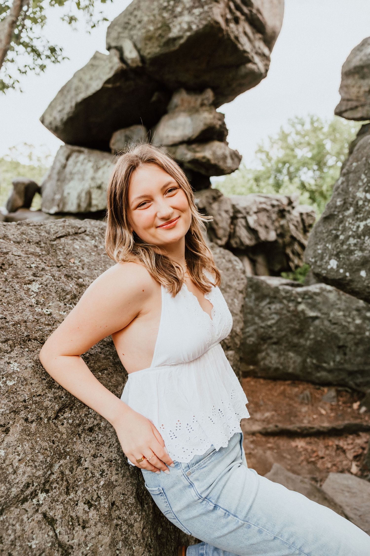 Smiling young woman in white top leaning against large rocks in a natural outdoor setting.